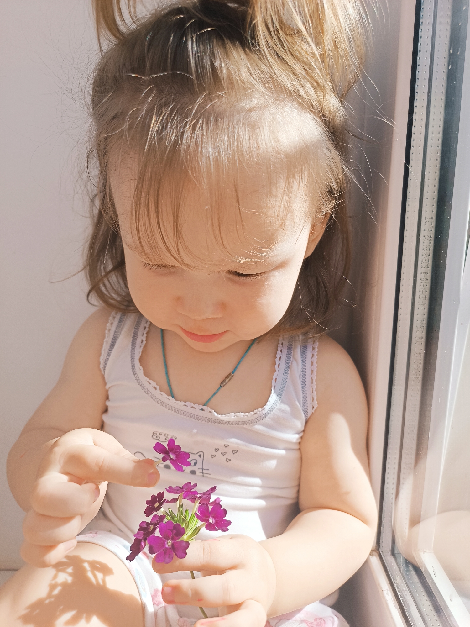 A young child with light hair in a ponytail is sitting indoors, close to a window, and holding a small bunch of purple flowers. The child is wearing a white top with a patterned trim and a blue necklace. The child is looking down at the flowers, examining them. The location cannot be verified to be in Izhevsk, Russia.