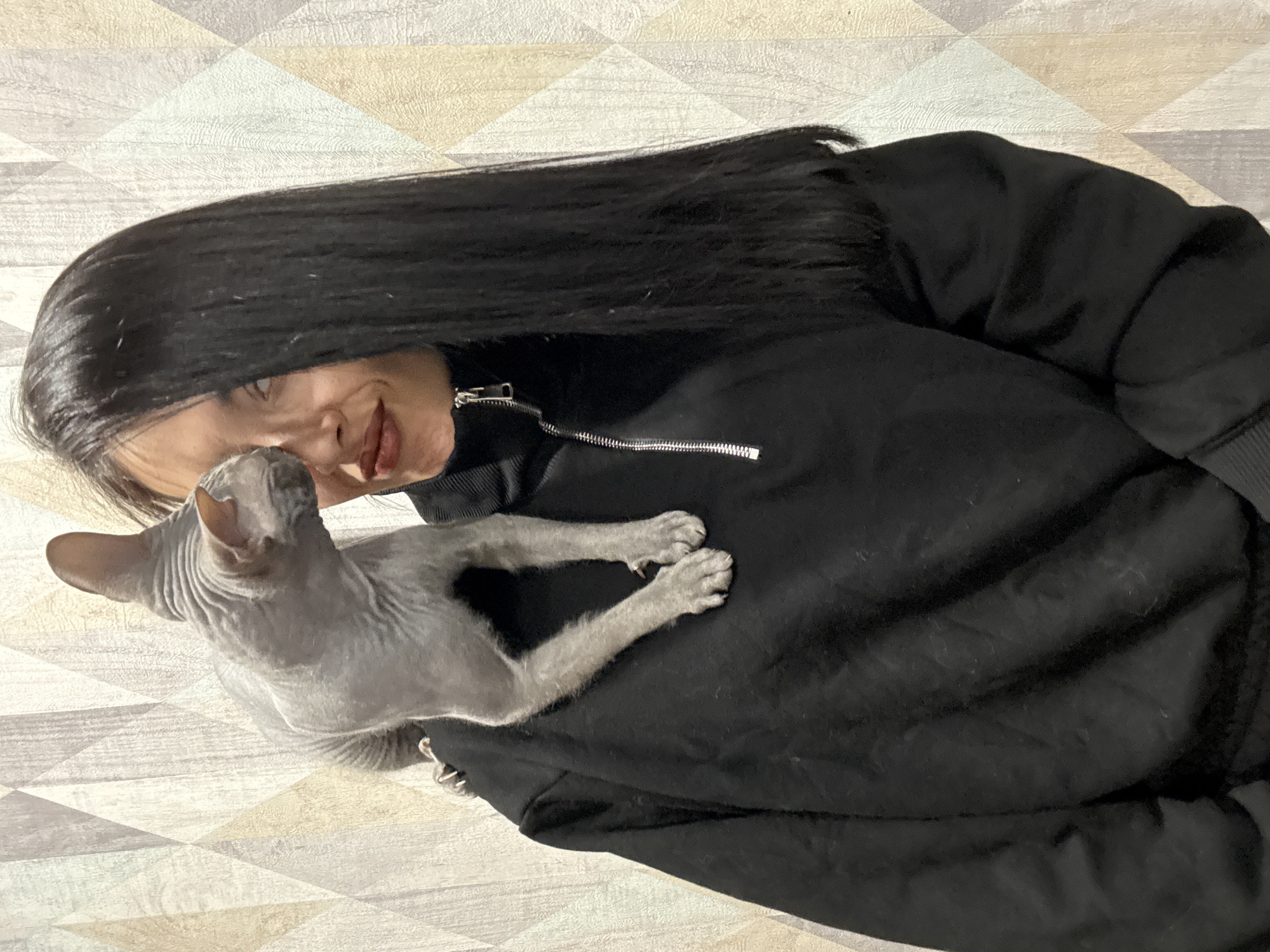 A woman with long black hair is holding a Sphynx cat against her black zip-up jacket. The woman is smiling, looking at the cat, and is indoors. The background shows a geometrically patterned surface. There is no evidence in the image to suggest the location is Votkinsk, Russia.
