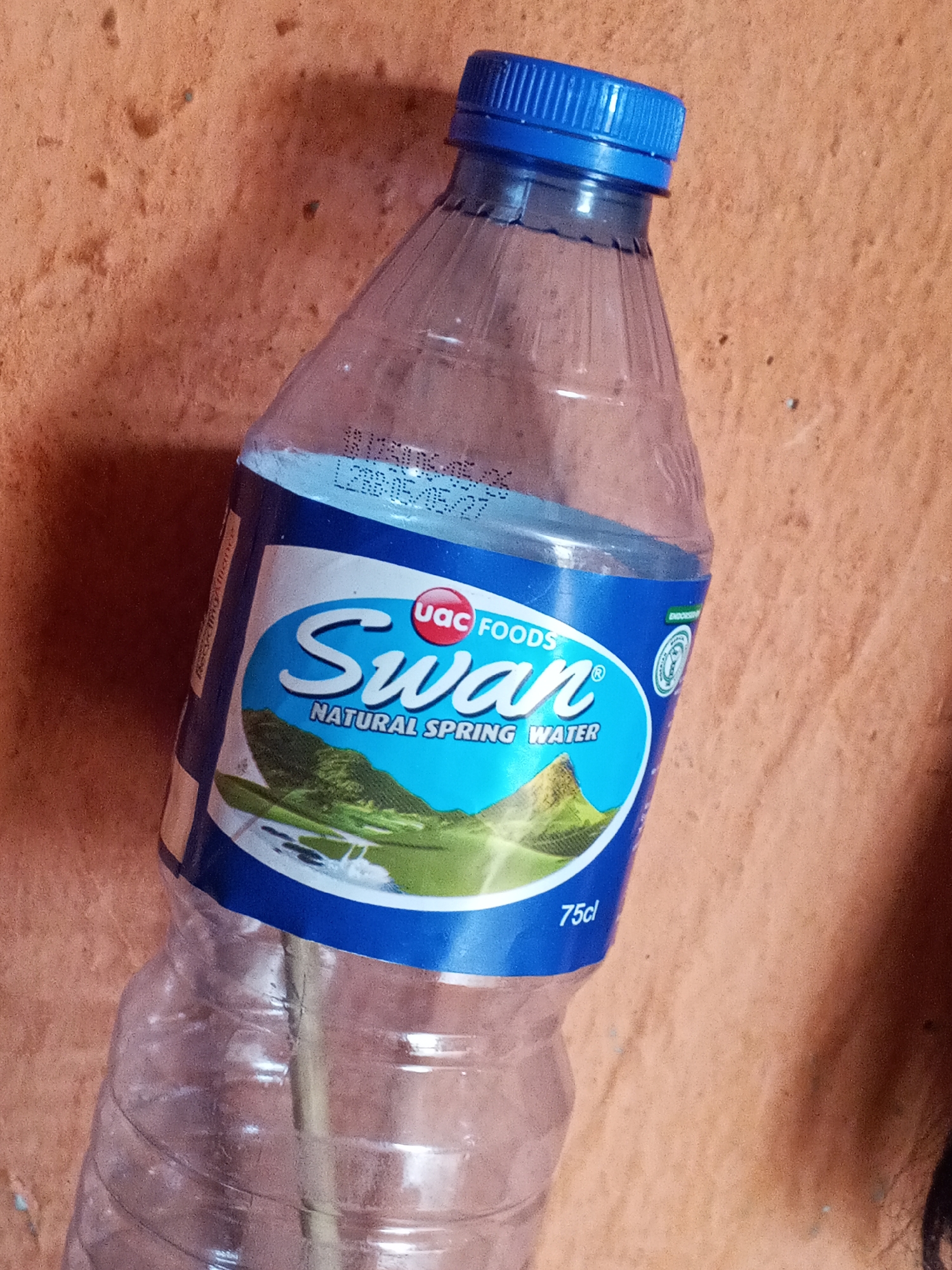 An empty 75cl plastic water bottle is positioned in front of a textured, light orange background. The bottle is branded "Swan NATURAL SPRING WATER" with a blue label. The label includes the phrase "UAC FOODS", and the bottle has a blue cap. The bottle’s expiration date is "L2BB-05/05/27". The bottle is clear and empty. Based on the branding, the bottle appears to be from Nigeria. The exact location in Gwong, Nigeria cannot be determined.