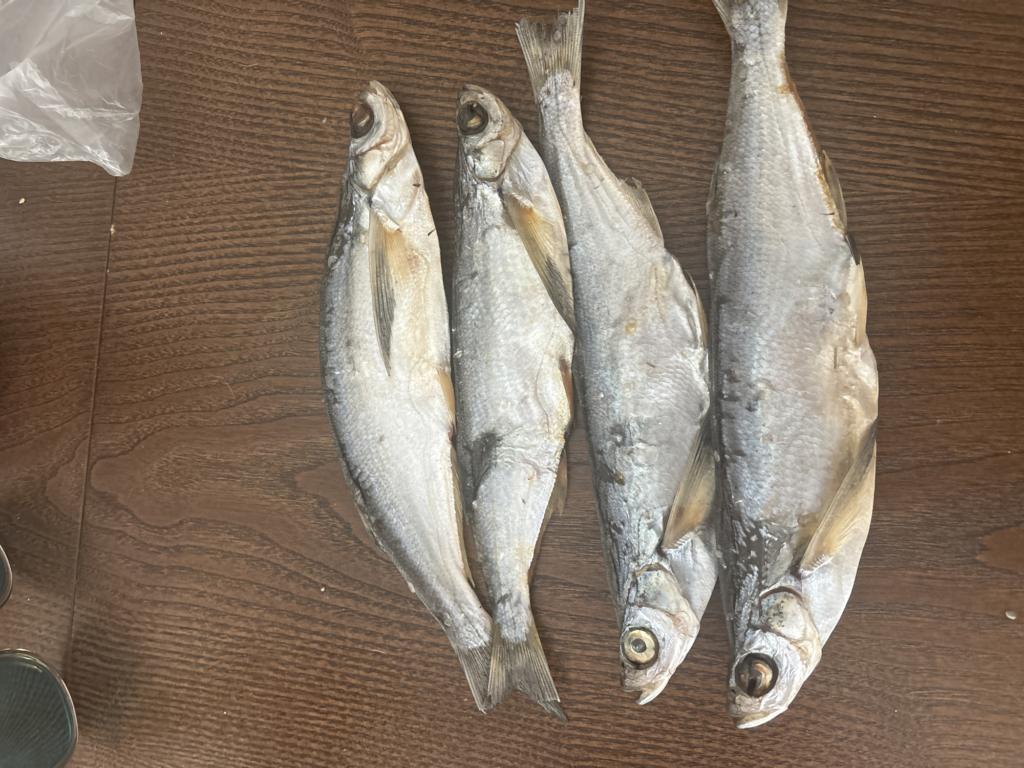 Four dried fish are arranged diagonally on a wooden surface. The fish are oriented head-to-tail, with their heads pointing towards the bottom left of the image. The wooden surface appears to be a table with a dark brown wood grain pattern. A crumpled white plastic bag is visible in the upper right corner of the image. There is no indication of the location in Chaikovsky, Russia.