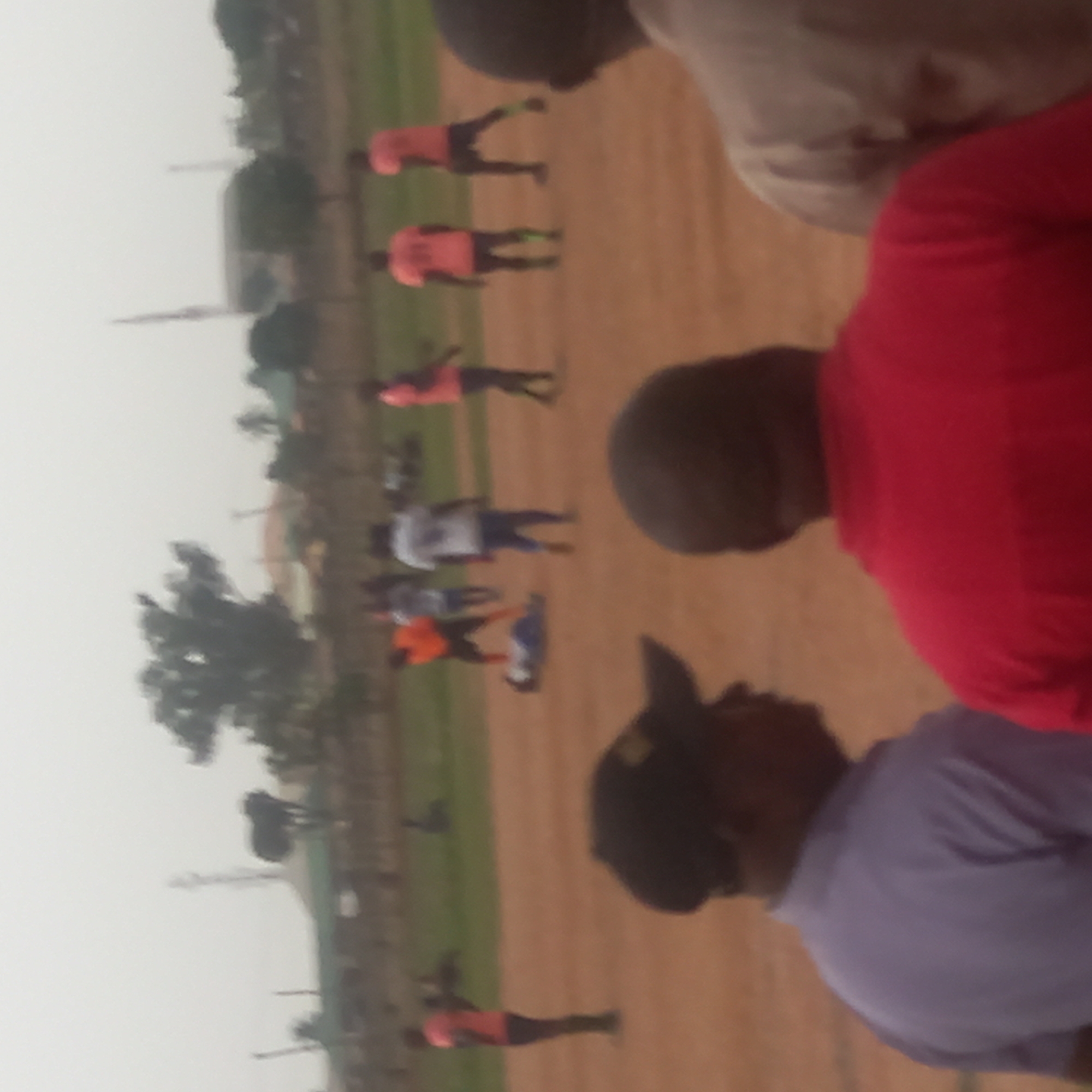 The image shows a group of people on a field in Narayi, Nigeria. Some people are standing or walking, and appear to be wearing sports clothing. Others are standing closer to the camera, and are wearing casual clothing. The field appears to be dirt, with a patch of grass. In the background are trees, buildings, and a tall thin structure. The weather appears overcast.
