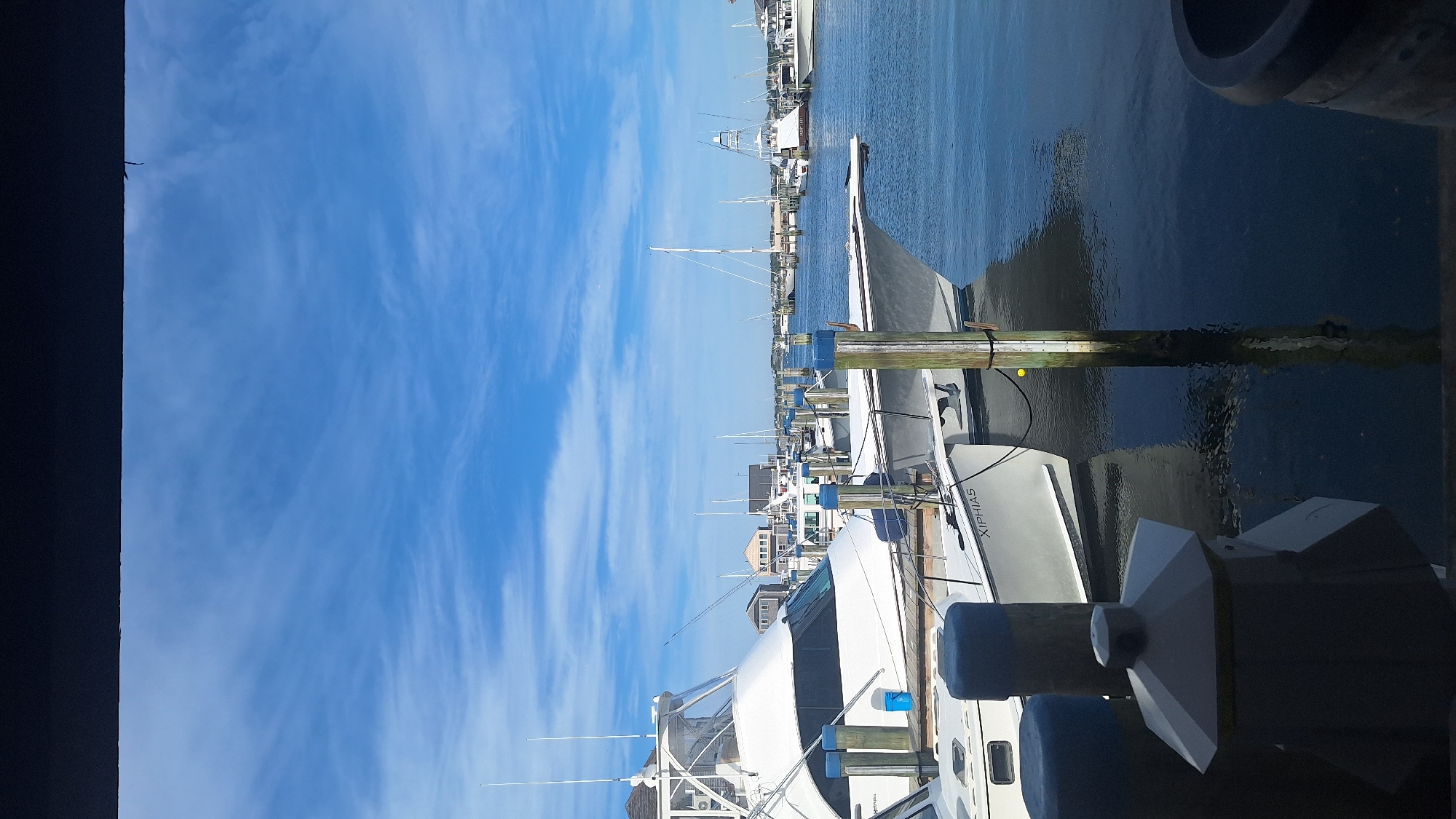The image depicts a marina in Nantucket, USA, under a bright, partly cloudy blue sky. Several white yachts and sailboats are docked side-by-side in calm blue water. In the foreground, two wooden pilings with blue caps are visible, along with a grey structure that appears to be a bollard or a decorative element. A white boat with the name "XIPHIAS" is prominently featured. In the background, more boats and buildings are visible, suggesting a bustling harbor. The water reflects the sky and the boats, creating a serene atmosphere.