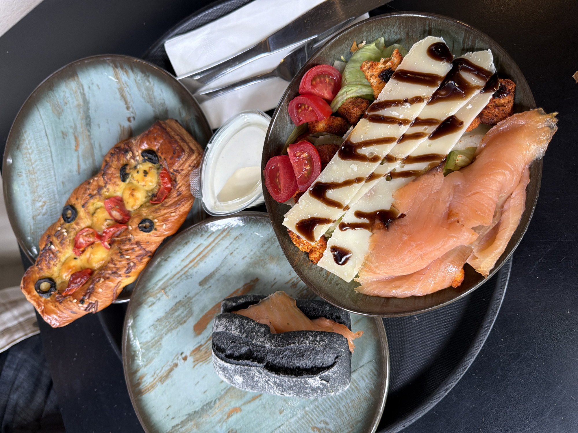 The image shows a tray of food, including a salad, pastry, and black bread sandwich. The salad contains lettuce, tomatoes, croutons, cheese with balsamic glaze, and smoked salmon. The pastry has cheese, tomatoes, and olives on top. The black bread sandwich also contains smoked salmon. A container of sauce and cutlery are visible. The location is likely a cafe or restaurant in Tbilisi, Georgia, based on the style of food and serving dishes.