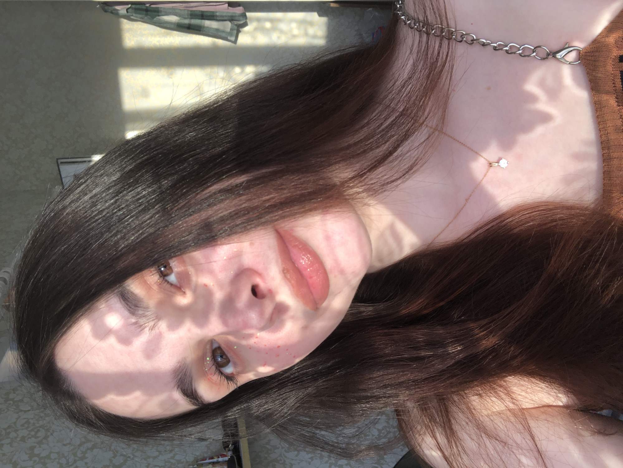 A young woman with long, dark brown hair is depicted in a close-up, tilted portrait. She is fair-skinned and wearing a brown top. She also has a silver chain around her neck and a delicate gold necklace with a small pendant. Light and shadow patterns from foliage fall across her face. Her lips are glossy, and she has some glittery makeup around her eyes and some red spots on her face, likely freckles.
The background shows a glimpse of a room with a window casting light, suggesting an indoor setting. The location is unspecified, so the city of Noviy, Russia, cannot be confirmed.