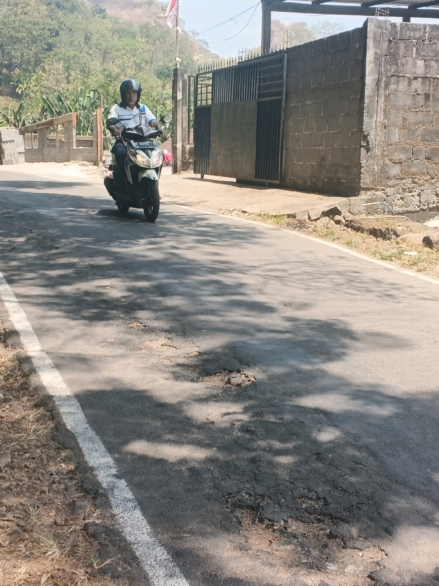 A man rides a motorcycle down a paved road in Leyangan, Indonesia. He is wearing a helmet, a white shirt with blue stripes, and dark pants. The motorcycle is white and black. The road is cracked and weathered, with a white line painted on the left side. Shadows of trees are cast across the road. In the background, there is a wall made of cinder blocks and a metal gate. Beyond that, a hillside covered in vegetation is visible. A red and white flag flies on a pole. The sky is clear and blue.