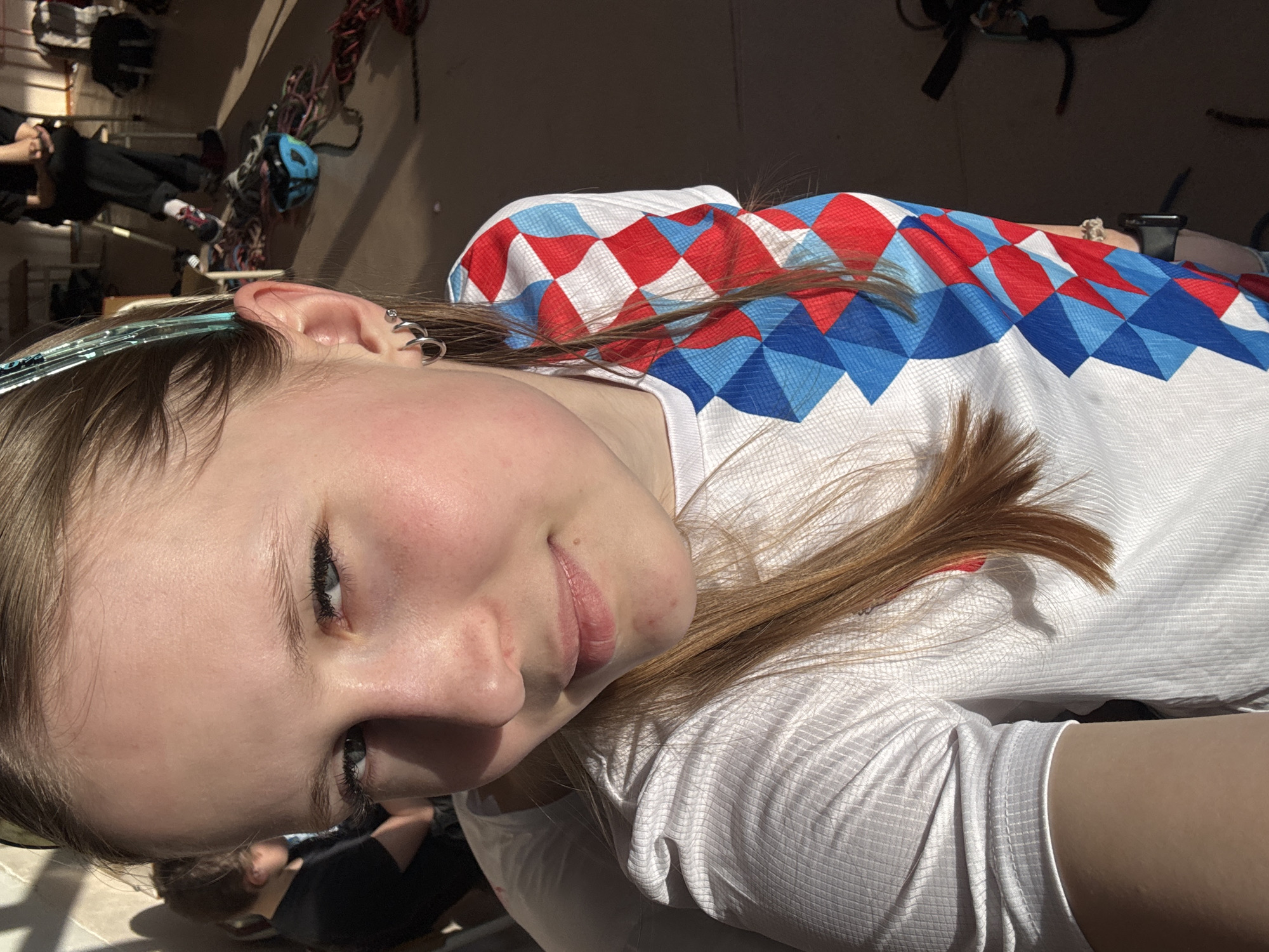 A fair-skinned person with blue eyes and light brown hair is centered in the frame. They are wearing a white shirt with a red and blue diamond pattern on the upper portion and a light blue headband. Multiple earrings adorn the left ear.
In the blurry background, partial figures of other people and bicycles are visible. The setting appears to be indoors with sunlight streaming in. There is no verifiable information regarding the location being Usolye, Russia.