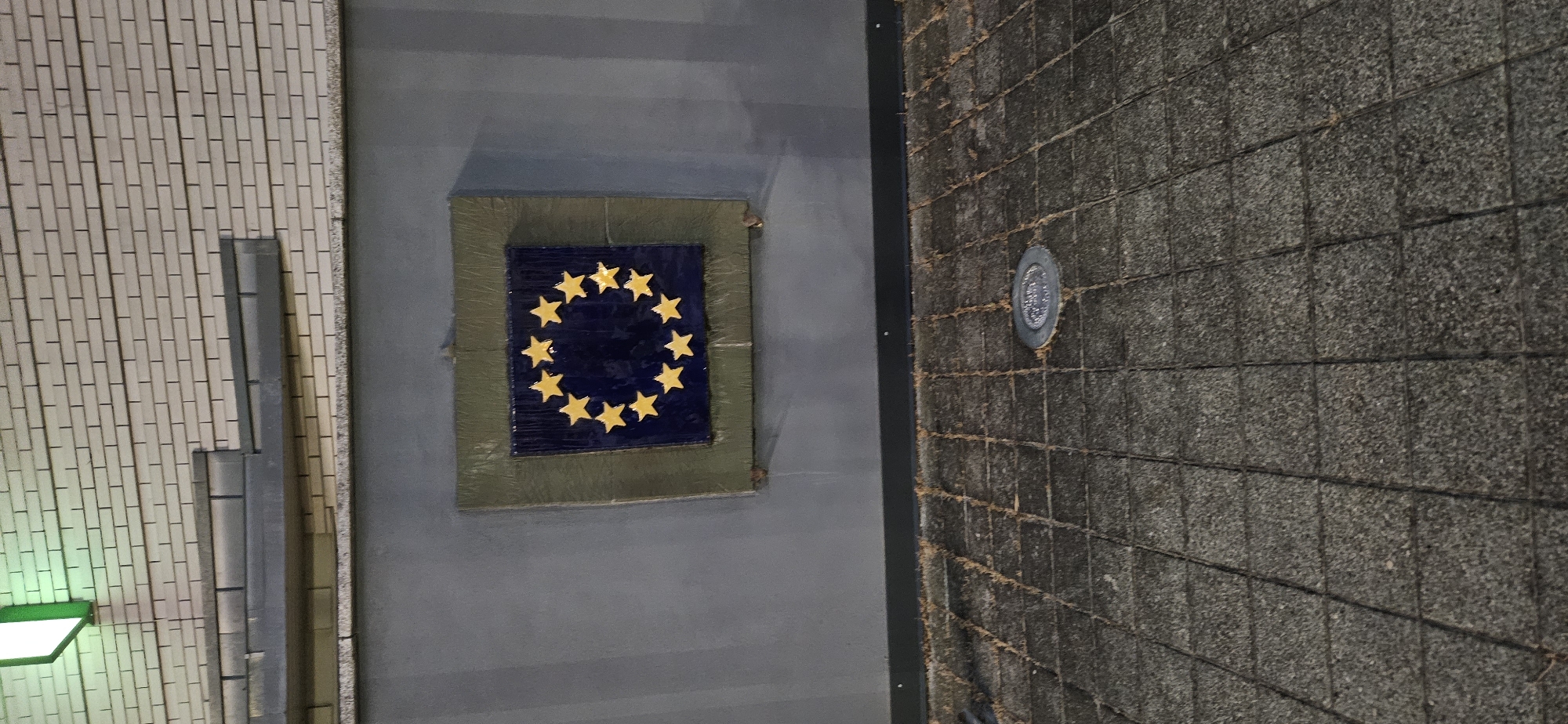The image shows a tiled facade with a square plaque featuring the European Union's flag. The flag has a blue background with a circle of 12 yellow stars. The plaque is mounted on a grey wall below a light-colored brick wall with horizontal grey decorative elements and a green illuminated sign. The foreground is a cobblestone pavement with a circular metal fixture embedded in it. The scene appears to be an outdoor public space, likely in the city of Hévíz, Hungary.
