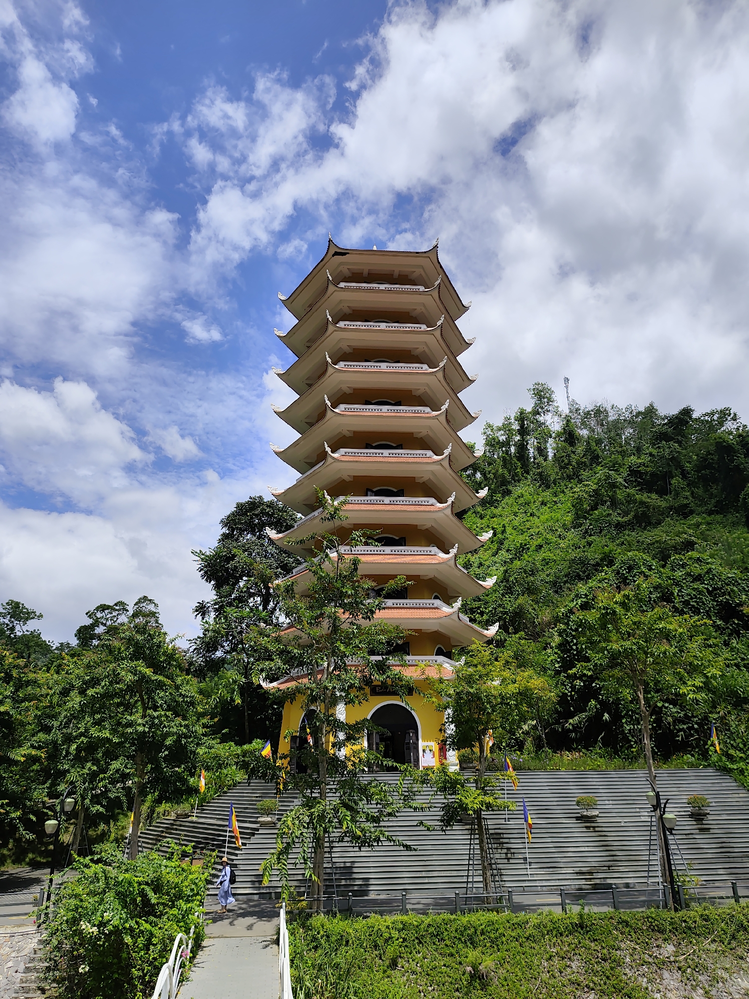 A seven-tiered pagoda with a yellow base and orange roof is centrally located in the image, set against a backdrop of lush green trees and a bright blue sky filled with white, fluffy clouds. The pagoda's tiered roof features distinctive upturned eaves. In front of the pagoda, a wide set of stairs leads up to the entrance, adorned with several colorful flags. A person in a light blue robe and a straw hat is walking up these stairs. The surrounding environment is dominated by dense foliage, with trees of varying sizes and a steep, vegetated hillside behind the structure. The scene appears to be a temple or a place of worship situated in a natural, hilly landscape.