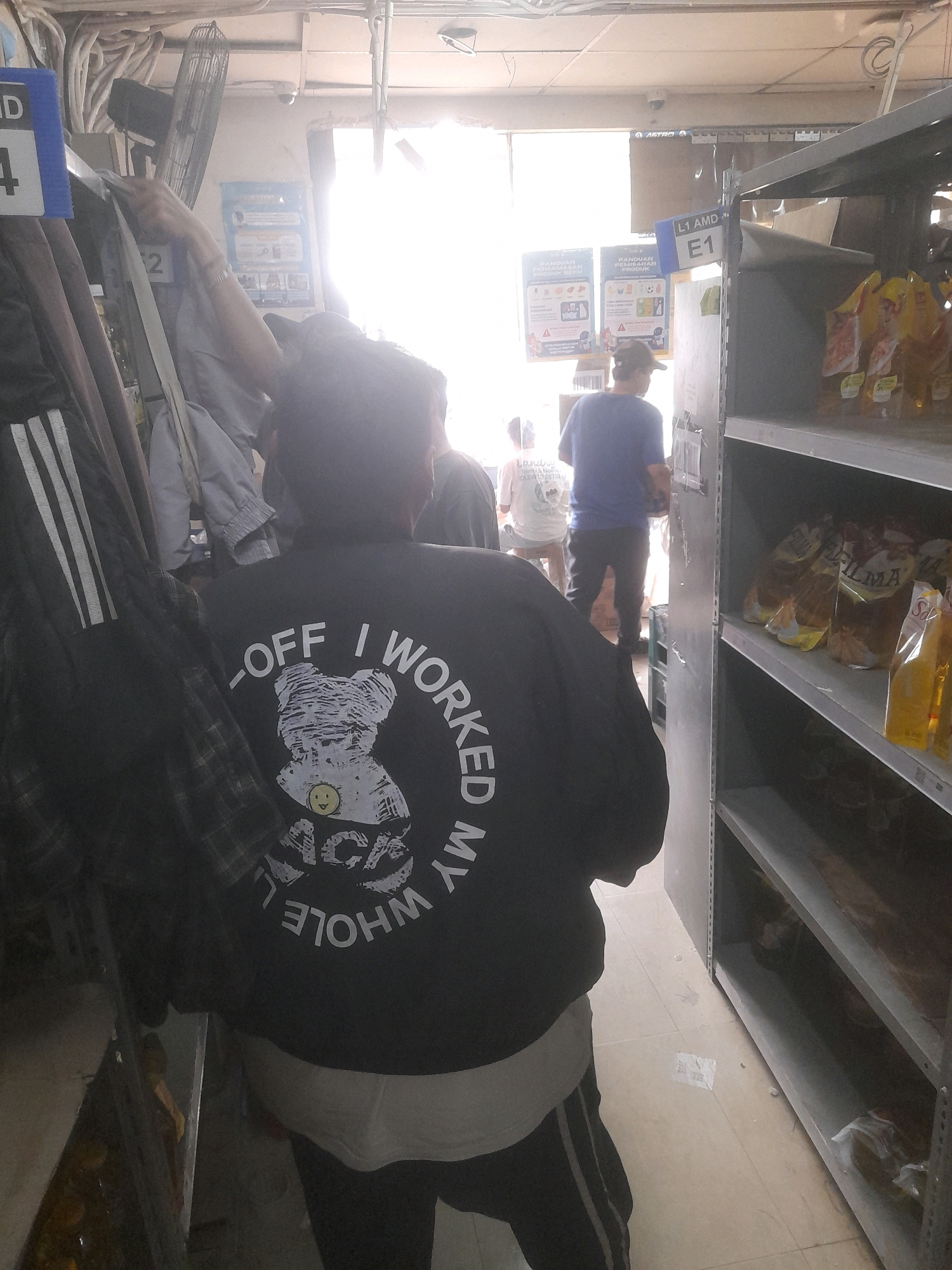 The image depicts an indoor scene, possibly a storage area or a retail backroom, with shelves stocked with various items. The environment appears cluttered, with electrical conduits and fans visible on the ceiling and walls. A bright light source, likely natural light from an opening, illuminates the background.
In the foreground, a person is seen from behind, wearing a black jacket with white text and graphics on the back. The text reads "-OFF I WORKED MY WHOLE." A graphic of a teddy bear wearing a smiley face and sunglasses is also present. The person is standing in a narrow aisle next to shelves.
Other individuals are partially visible in the background. One person is wearing a blue shirt and dark pants, standing near a shelf. Another person is wearing a white t-shirt.
Several signs are visible on the wall and shelves. One sign reads "L1 AMD E1". Another sign shows product information and instructions. There are also various items on the shelves, including what appear to be food products in packaging. The floor is tiled. The scene suggests a utilitarian, possibly industrial or commercial, setting in Bekasi, Indonesia.