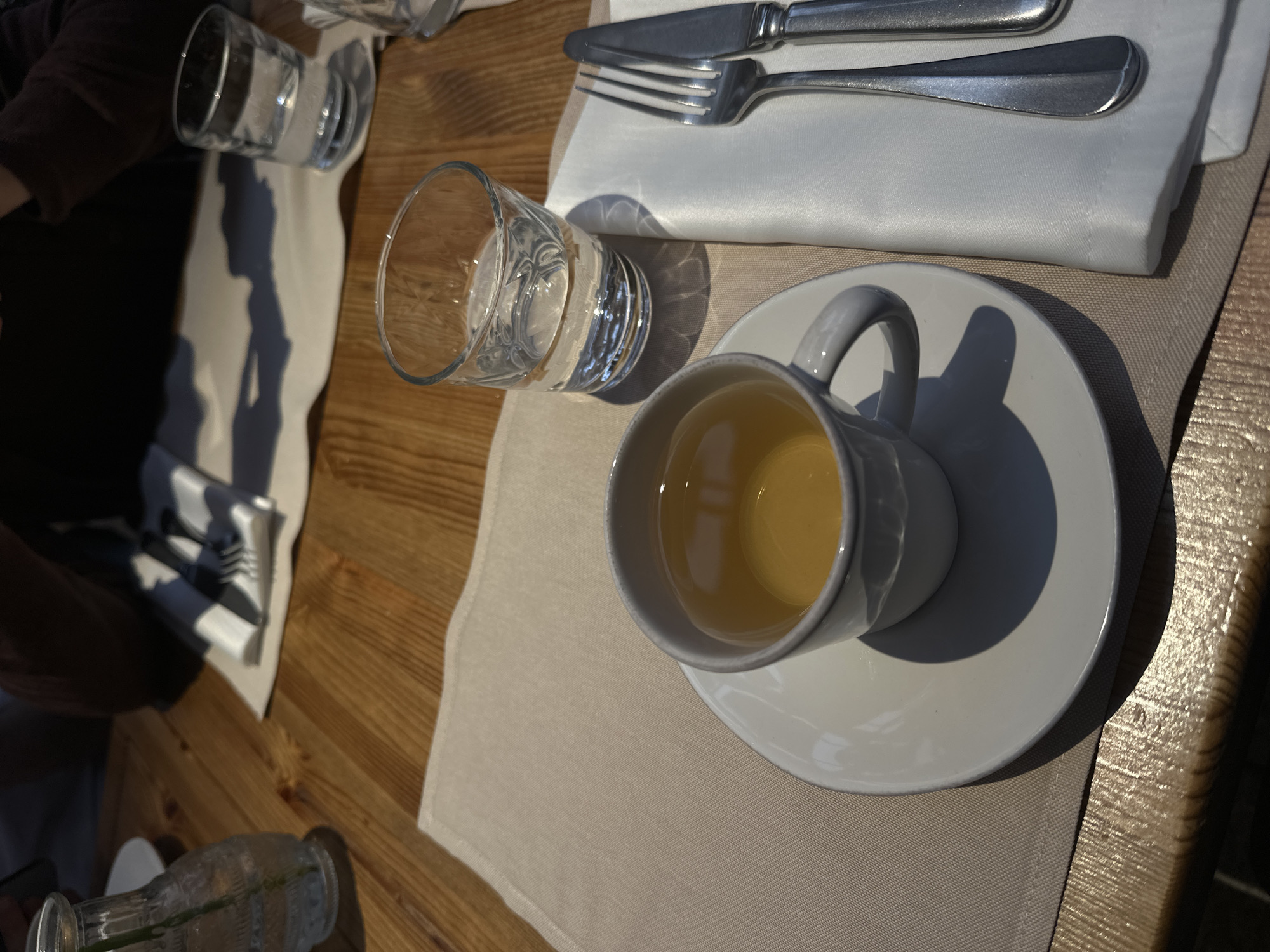 The image shows a table setting with a cup of tea on a saucer, a glass of water, silverware on a napkin, and another glass of water in the background. The table is wooden with a placemat. The location is not specified and cannot be determined from the image. The scene appears to be indoors or in a shaded outdoor area, given the lighting.