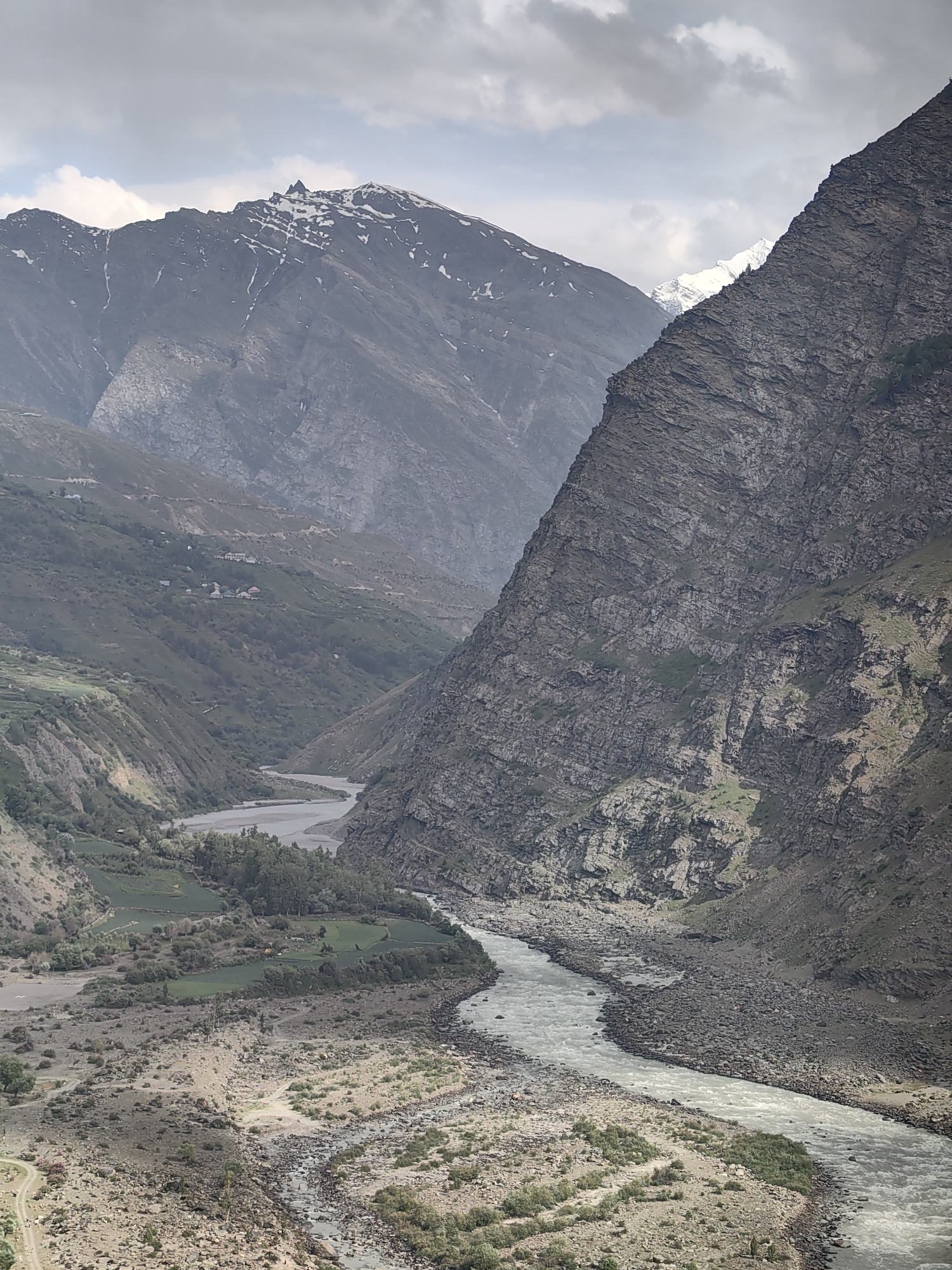 The image shows a rugged, mountainous landscape with a wide, winding river flowing through a valley. Steep, rocky slopes dominate the foreground and mid-ground, with a sheer, dark gray cliff face on the right. In the distance, higher mountains are visible, some with patches of snow. The valley floor features patches of green vegetation and what appear to be small, scattered settlements or farmsteads. The river is milky-white, indicating it may be carrying sediment. The sky is overcast with diffused light. The scene depicts a remote, natural environment. The location is stated to be Kirting, India.