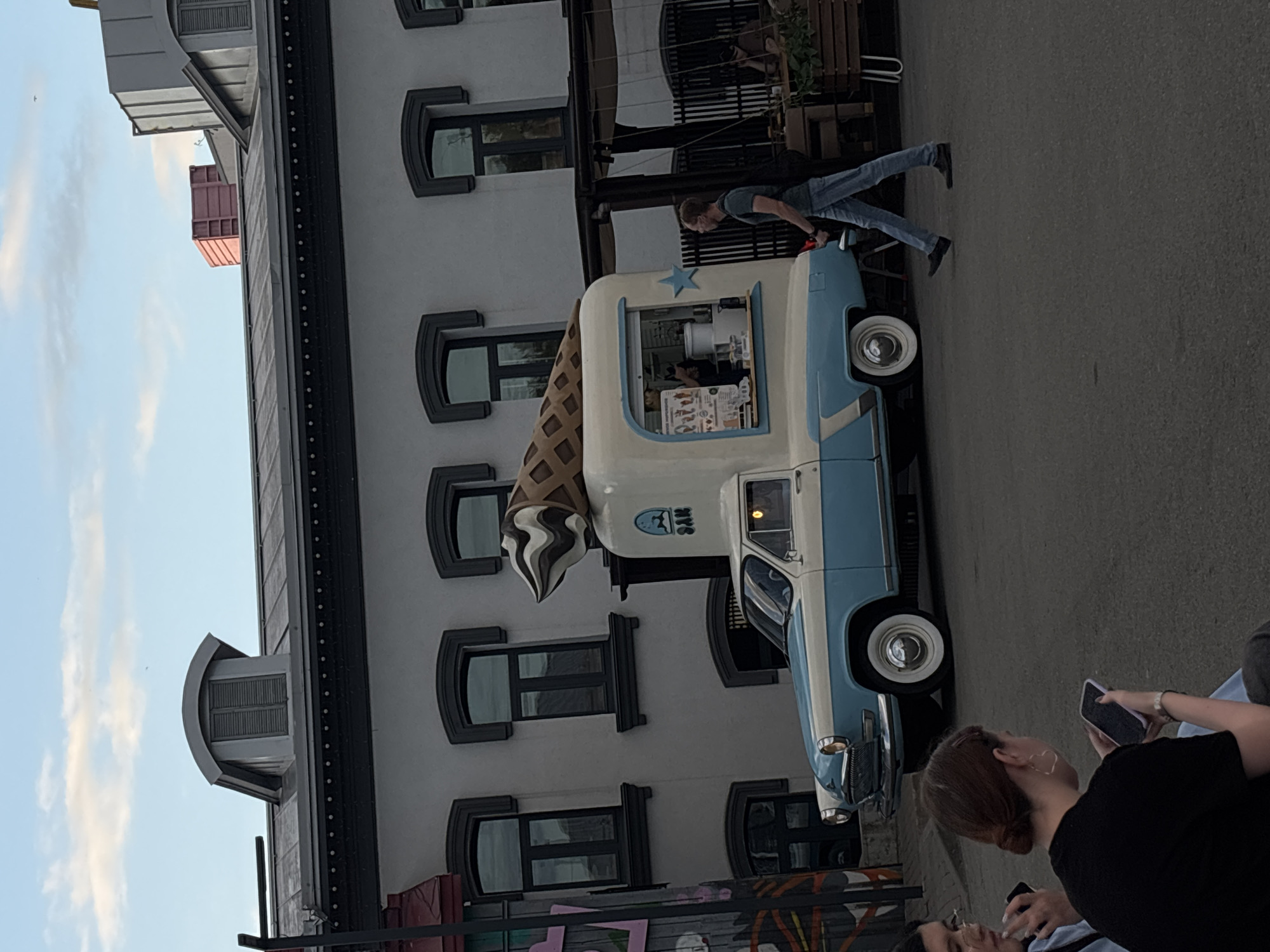 A vintage light blue and white ice cream truck shaped like a giant ice cream cone is parked on a paved surface in front of a gray building with arched windows. A man in jeans and a dark t-shirt is walking past the truck. Two people, a woman in a black shirt and a woman with glasses, are seated in the foreground, looking at their phones. The building has a dark roofline with decorative trim and several dormer windows. There is a covered outdoor seating area to the right of the truck. The sky is cloudy. The image appears to be taken in the city of Ufa, Russia, possibly in a public space or market area.