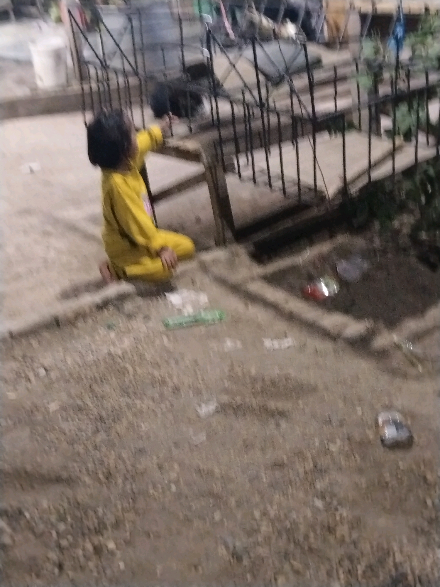 A young child, appearing to be of Southeast Asian descent, is squatting on a dirt and concrete surface. The child is wearing a bright yellow hooded jacket and pants. The child's back is to the camera, and they are reaching out towards a black and white dog that is partially visible behind a metal fence. The fence appears to be part of a small enclosure. In the background, there are some indistinct objects and structures, possibly part of an outdoor area in a residential or urban setting. Scattered on the ground are some pieces of debris and litter. The scene is dimly lit, suggesting it might be evening or an overcast day. The location is described as the city of Suka Ramai, Indonesia.