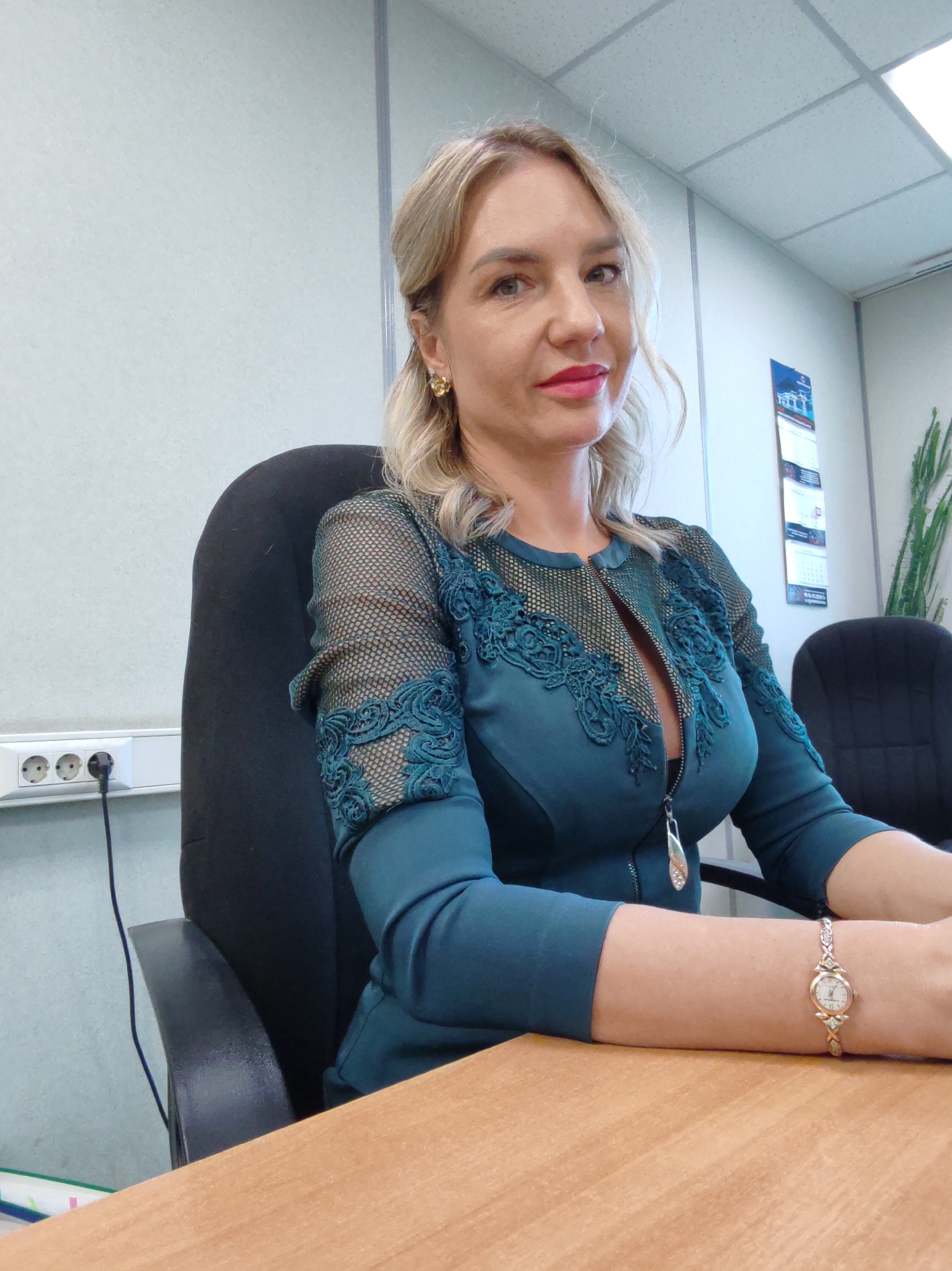 A woman is seated at a wooden desk in an office. She is wearing a teal-colored dress with intricate lace detailing on the sleeves and neckline. Her blonde hair is styled in soft waves. She is wearing makeup, including bright pink lipstick and earrings. On her left wrist, she wears a delicate gold watch with a white face. She is looking towards the camera with a slight smile. In the background, there are office walls, a ceiling with fluorescent lighting, a power strip with three outlets, and two black office chairs. A tall green plant is visible on the right side of the image. A calendar is hanging on the wall behind her. The overall impression is of a professional setting.