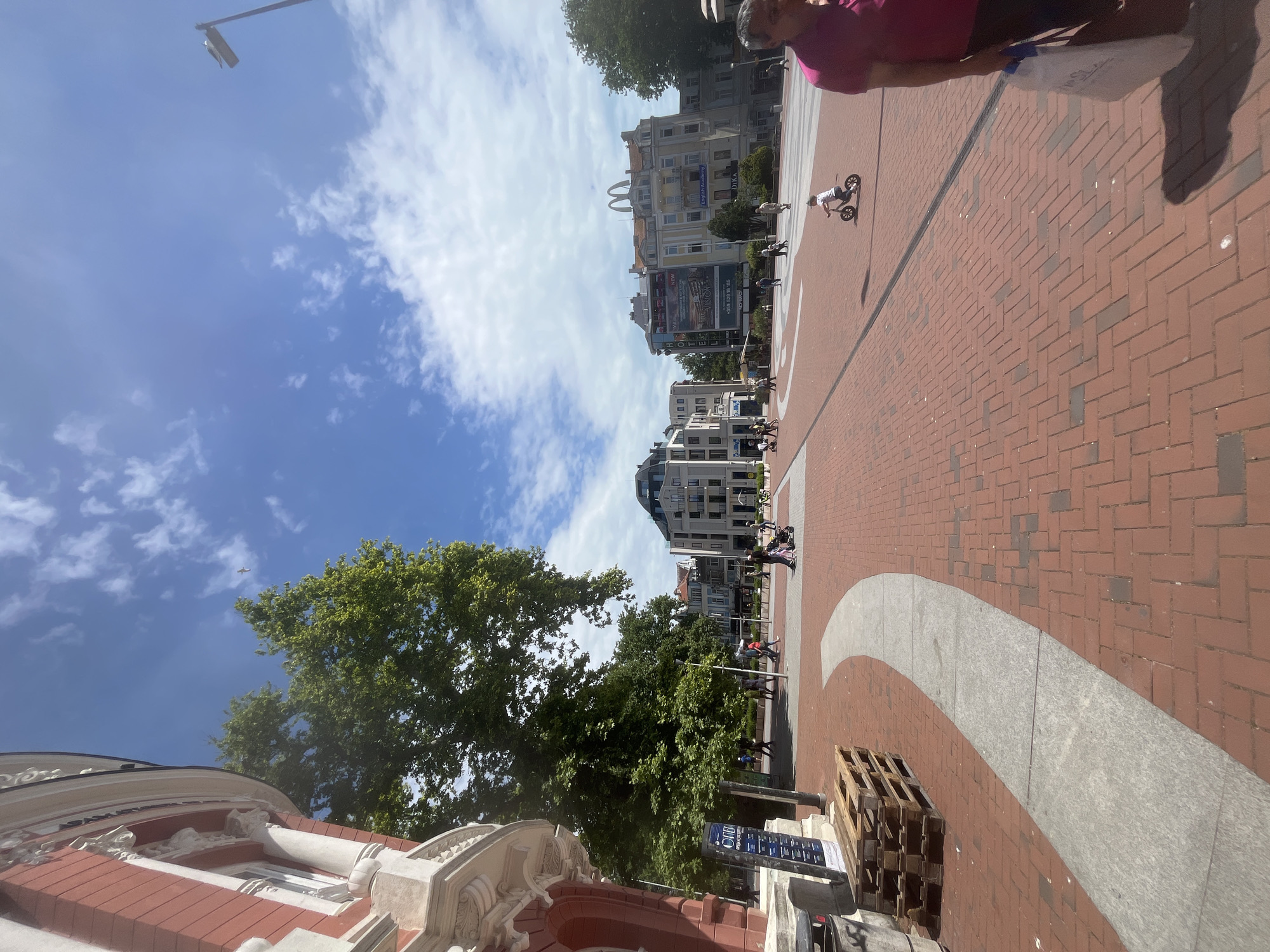 The image shows a wide brick-paved plaza in Varna, Bulgaria, on a sunny day with a blue sky and scattered clouds. The plaza has a curved gray stone design embedded in the brick. People are walking and a child is riding a scooter on the plaza. A building with a red and white façade is visible on one side of the plaza, partially obscured by trees. In the background, there are several buildings of varying heights and architectural styles. A street lamp stands tall in the distance. Wood pallets are stacked on the plaza's edge.