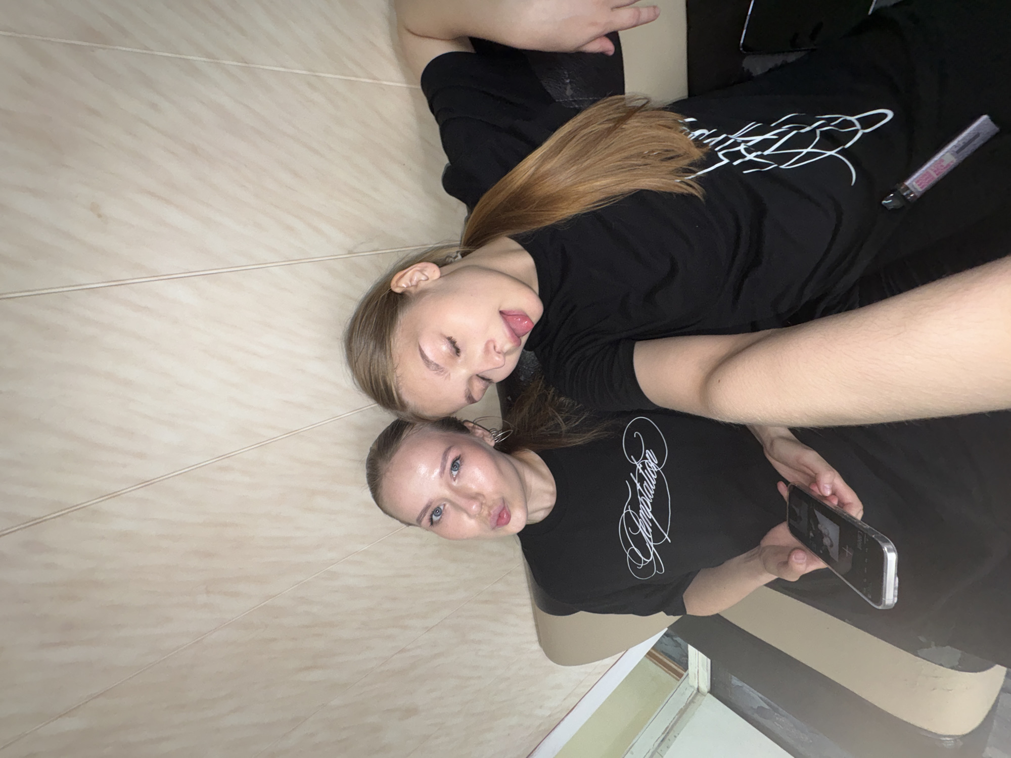 Two young women are captured in a casual, intimate shot, likely a selfie or self-timer photograph, as they lie down on what appears to be a light-colored tiled floor or surface. The image is oriented with the subjects lying horizontally across the frame.
The woman in the foreground is looking directly at the camera with a slight pout, her blue eyes wide open. Her light brown hair is pulled back, and she is wearing a black t-shirt featuring the word "Temptation" in an ornate white script. She holds a smartphone in her hands, its screen visible but details unclear.
Beside her, the second woman is lying down with a playful expression, sticking out her tongue and having her eyes closed or partially closed. Her long, light brown hair is spread out behind her head. She also wears a black t-shirt, which has a distinct white abstract or stylized graphic design on it. A slim, rectangular object, possibly a vape pen or cosmetic item, with some illegible pink branding, rests near her on the dark surface beside the tiled area.
The background consists primarily of the light-colored, possibly beige or cream, tiled surface with visible grout lines. A darker surface, likely a bed or sofa, is positioned next to the tiled area. In the lower right corner, a partial view of what appears to be a window or doorway with a dark frame and some lighter interior is visible. The overall mood of the image is relaxed and friendly. The scene is set indoors, with the context given as Chaikovsky, Russia.