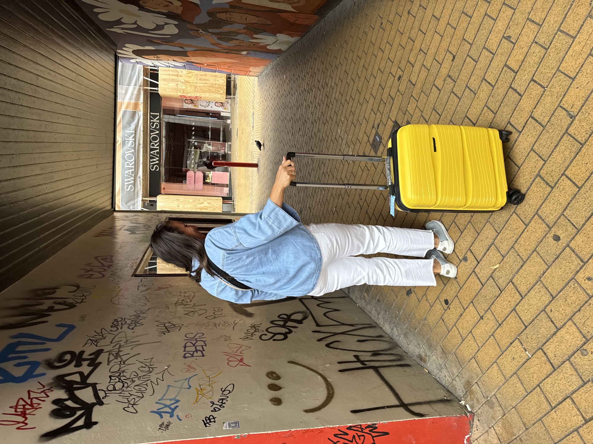 A person is walking through a narrow alleyway with a yellow suitcase. The walls of the alley are covered in graffiti, and the ground is paved with yellow bricks. In the background, there is a Swarovski store. The alley appears to be located in the city of Groningen, Netherlands. The person is wearing a blue denim shirt, white pants, and gray shoes. They are holding the handle of the yellow suitcase, which has a black trim and four wheels. A small black bird is standing on the ground in front of the Swarovski store. The Swarovski store has large windows and a sign that says "SWAROVSKI" in silver letters. The store is boarded up with wood. The walls of the alley are decorated with murals on the right side.