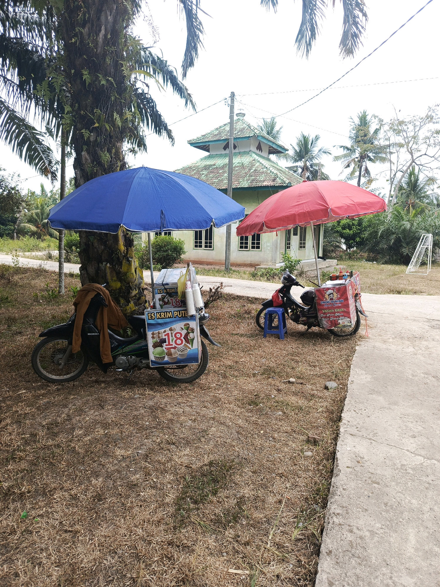 In Megang Sakti V, Indonesia, two motorcycles serve as mobile food carts under separate umbrellas. The motorcycle on the left, under a blue umbrella, displays signage for "ES KRIM PUTIH" (white ice cream) and features an image of ice cream cones with the number "18". A brown jacket is draped over the handlebars. The motorcycle on the right, under a red umbrella, has a food cart with signage for "CIRENG ISI AYAM" (stuffed chicken cireng) and mentions "Berbagai Rasa" (various flavors) and "Cireng Pilihan Menu" (menu choice cireng). A blue stool is placed beside it. Both motorcycles are parked on dry, brown grass in front of a building with a green roof, surrounded by palm trees and other vegetation. A soccer goal is visible in the background.