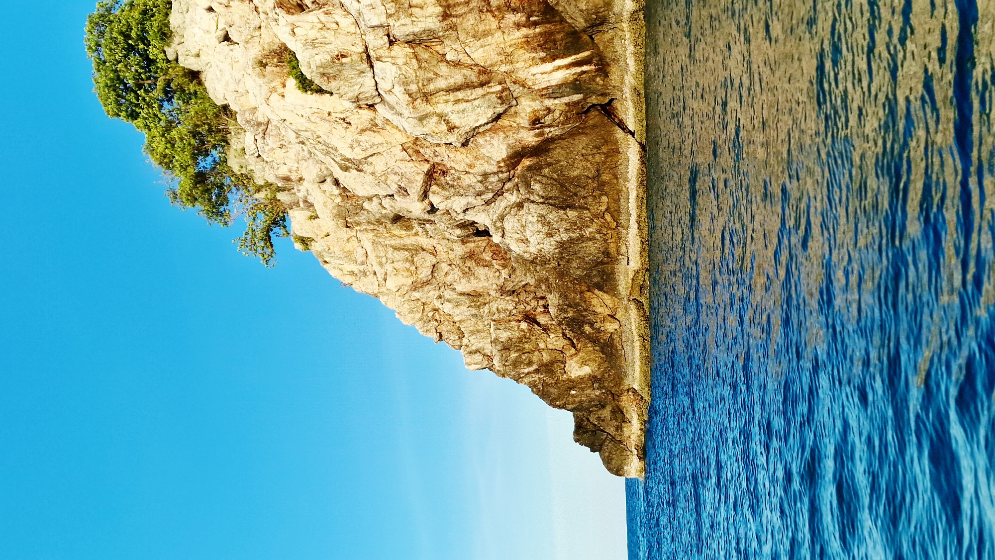 A vertical, full shot captures a rocky cliff face jutting out into bright blue water under a clear, cloudless sky. The cliff, composed of tan and brown rock, is partially covered by patches of green vegetation on its upper edge. The water in the foreground is a deep, vibrant blue, with slight ripples creating a textured surface that reflects the light and the cliff. The scene is devoid of people or man-made structures, presenting a natural coastal landscape. The lighting suggests a bright, sunny day.