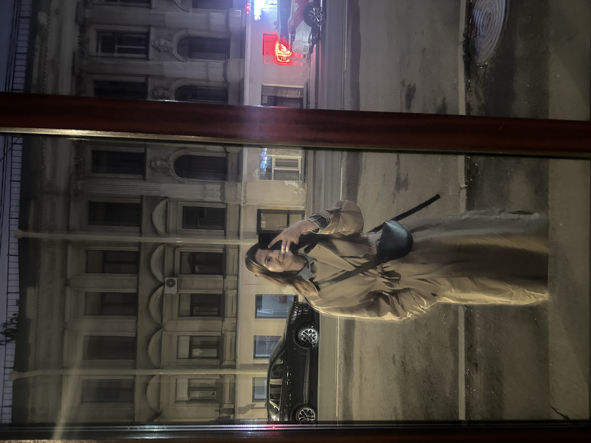 The image is a reflection of a woman taking a selfie in a window. The woman is wearing a tan trench coat and a blue denim shirt. She is holding her phone in her right hand and posing for the picture. Behind her is a reflection of a building with many windows. There is a black car parked on the street in front of the building. A red sign is visible on the building. The image appears to have been taken at night. The location is most likely the city of Saint Petersburg, Russia.