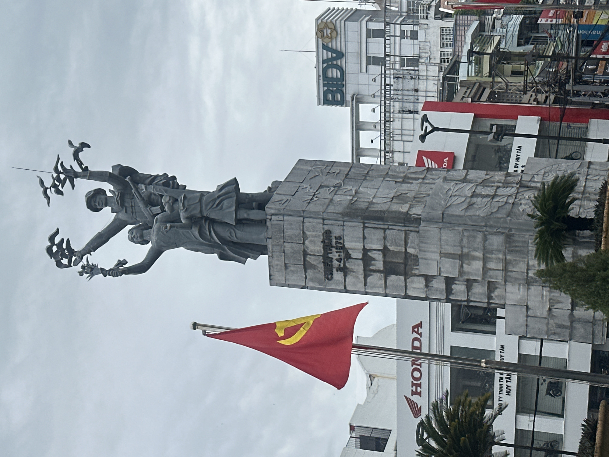 The image depicts a statue in Nha Trang Ward, Vietnam, with a red flag bearing a gold hammer and sickle in the foreground. The statue is of a group of people, possibly soldiers, holding up doves. Behind the statue, a building with "BIDV" signage is visible, along with other commercial establishments displaying "HONDA" signage. The environment suggests an urban setting. A date inscribed on the statue reads "2-4-1975" and a sign reads "CHIẾN THẮNG". There are palm trees. The sky is overcast.