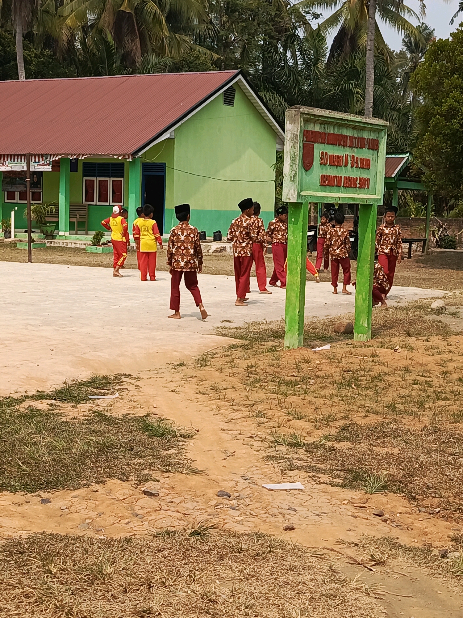 The image depicts an outdoor scene at what appears to be a school in Megang Sakti IV, Indonesia. In the foreground, dry grass and sandy soil are visible. A group of children, dressed in patterned shirts and maroon pants, are scattered across a concrete courtyard. Some are walking, while others are standing or crouching. Two children in the background wear yellow shirts and red pants. A light green building with a red roof stands behind the children, with several windows and an open doorway. To the right of the building is a sign with green posts and a weathered sign that reads "SD NEGERI 28 BUKANGA KECAMATAN BUKANGA BINTI". Palm trees and other greenery are present in the background. The overall impression is a sunny day at a rural school.