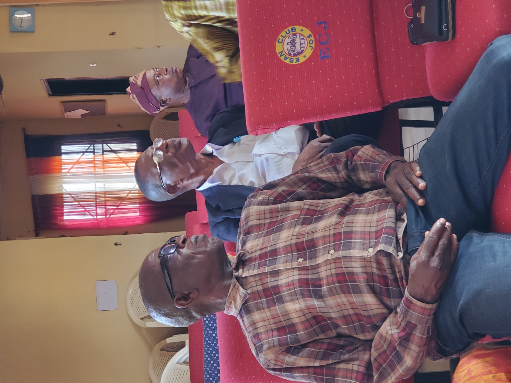 The image depicts three men sitting on red chairs. The man in the foreground is wearing a plaid shirt and jeans. The man in the middle is wearing a white shirt and a navy jacket. The man in the background is wearing a purple shirt and a patterned hat. The chairs have a logo on them that reads "CLUB ESAN". The location appears to be inside a room with a window in the background. It is not possible to confirm the exact location in Jos, Nigeria, from the given image.