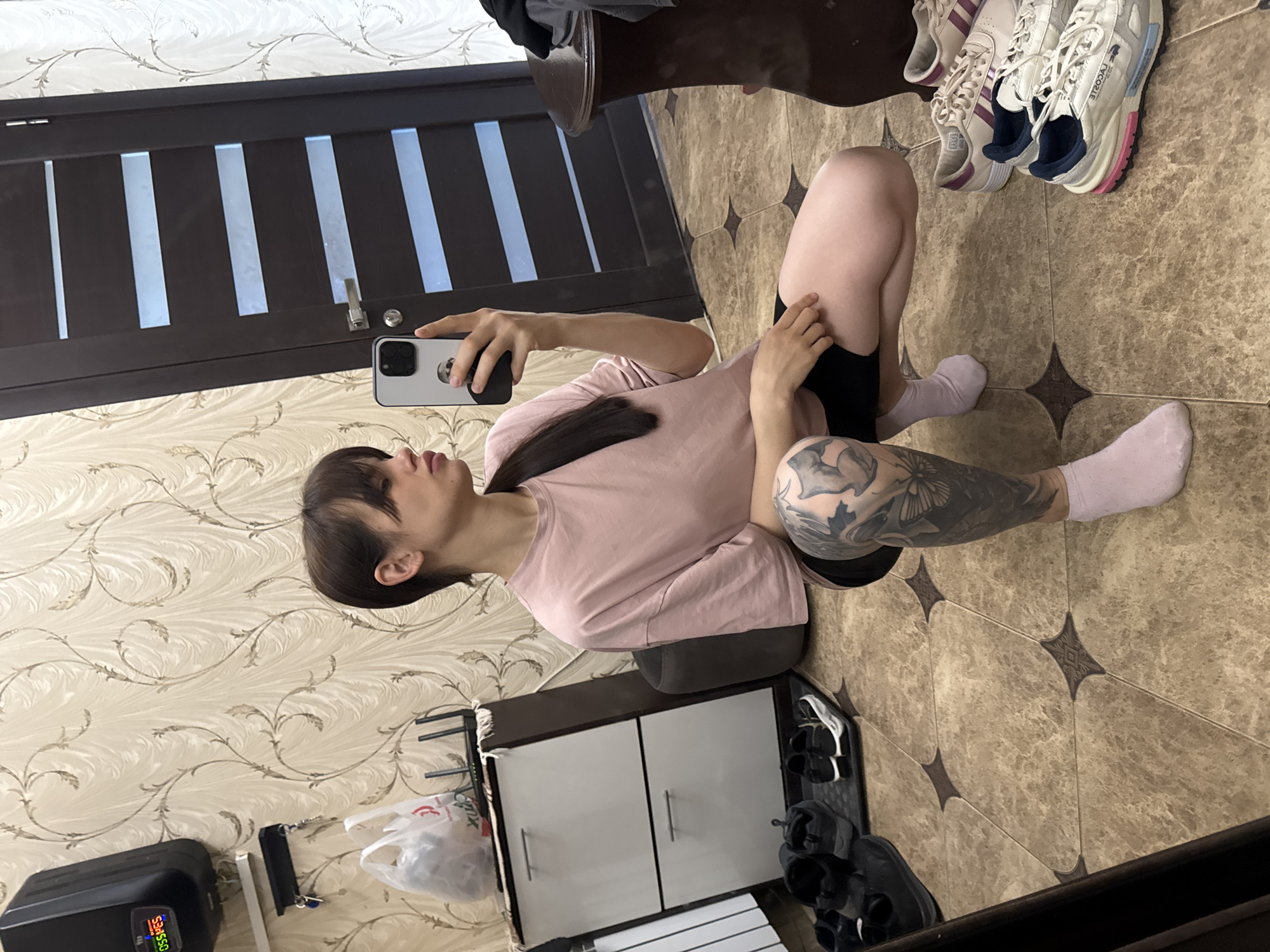A person is seated indoors, holding a smartphone, likely taking a selfie in front of a mirror. The individual has dark hair, bangs, and is wearing a pink shirt, black shorts, and light pink socks. Visible on the person's leg is a tattoo. The setting appears to be a domestic interior. Behind the person, the background includes a door with vertical panels, patterned wallpaper, a shoe cabinet, and several pairs of shoes. The floor is tiled. The location of Rostov-on-Don, Russia cannot be verified with the image alone.