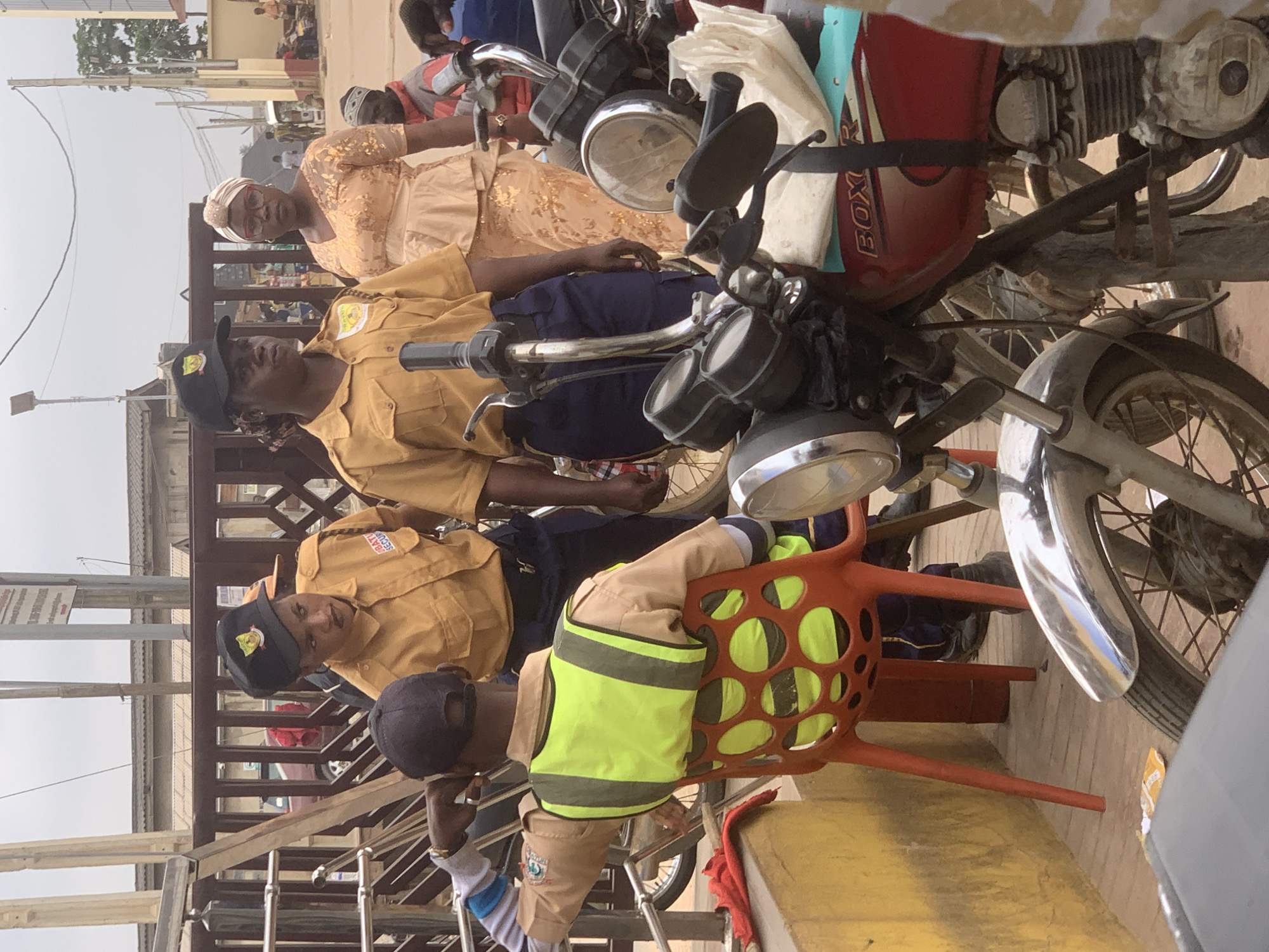In Shagamu, Nigeria, a group of individuals is gathered near a street with motorcycles. Two women in matching yellow security uniforms and black caps are conversing. One woman is looking to the right, while the other is looking to the left. A third person, seated on a bright orange chair and wearing a yellow security uniform with a high-visibility green vest, is facing away from the camera and leaning to their left, resting their elbow on something out of frame. Behind them, a woman in a peach-colored dress and headscarf stands looking towards the security personnel. In the foreground, parts of several motorcycles are visible, including handlebars, lights, and a red fuel tank with "BOXER" written on it. The ground is paved, and a yellow wall is visible on the left. The background shows buildings, a wooden fence, and other people in the distance. The sky is overcast.