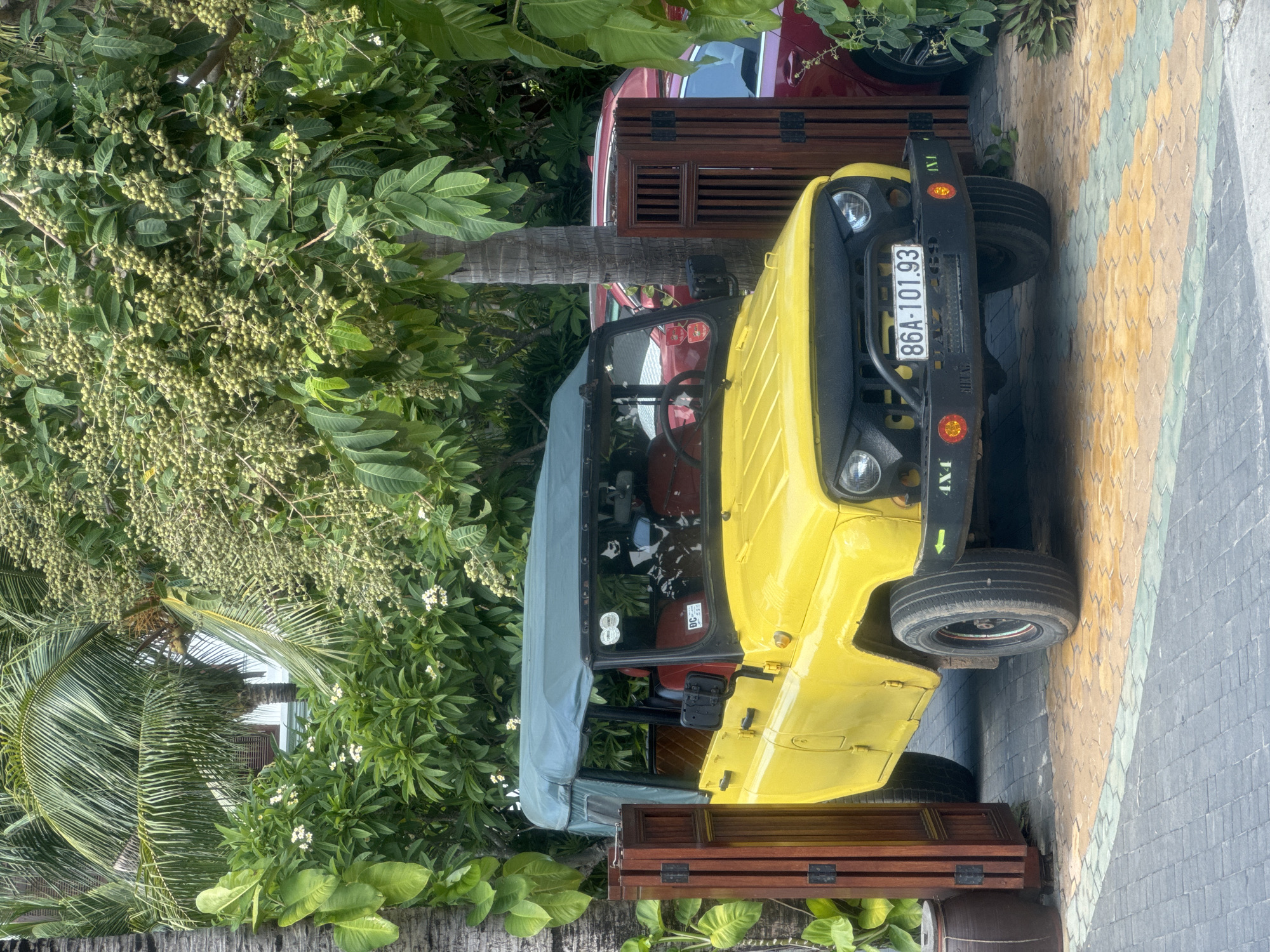 A yellow jeep with a black license plate numbered "86A-101.93" is parked on a paved surface near vegetation. The jeep is partially obscured by a wooden structure and a red car. The setting is Phu Thuy Ward, Vietnam.