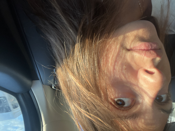 A young adult female is depicted in the interior of a vehicle in L'Haÿ-les-Roses, France. Her head is slightly turned, with her dark brown eyes gazing upwards and to the right. Her light brown hair, exhibiting auburn tones from direct sunlight, cascades over her shoulders and chest. Strong, direct sunlight illuminates the right side of her face and hair, casting distinct linear and mottled shadow patterns across her cheek and forehead. Her lips are closed in a neutral expression. A light-colored garment, possibly a white top, is visible on her lower left side. The vehicle's interior features a dark, likely black, leather seat or upholstery behind the subject. A light-colored car pillar and a dark window frame are visible on the right. Through the vehicle's window, a white utility vehicle, possibly a van, is discernible against a partly cloudy blue sky, indicating an outdoor, daytime setting.