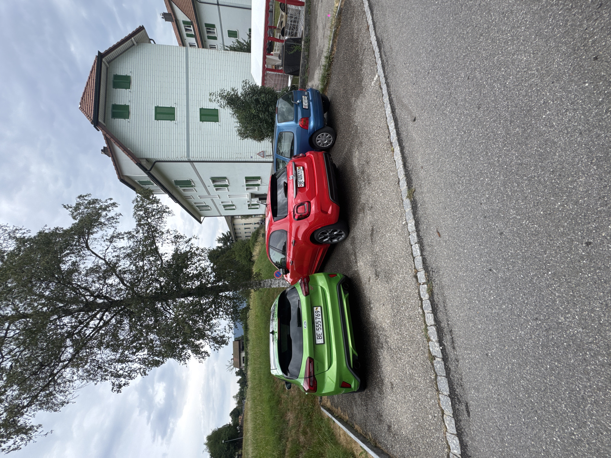 Three cars are parked in a gravel lot next to a paved road. The cars are, from left to right, a green Ford Focus, a red Fiat 500X, and a blue Renault Twingo. The license plates of the Ford and Fiat are visible. Behind the cars, a light blue apartment building with green shutters stands under a cloudy sky. A mature tree with dense foliage is positioned to the left of the cars. The environment appears to be a residential area in Tavannes, Switzerland.