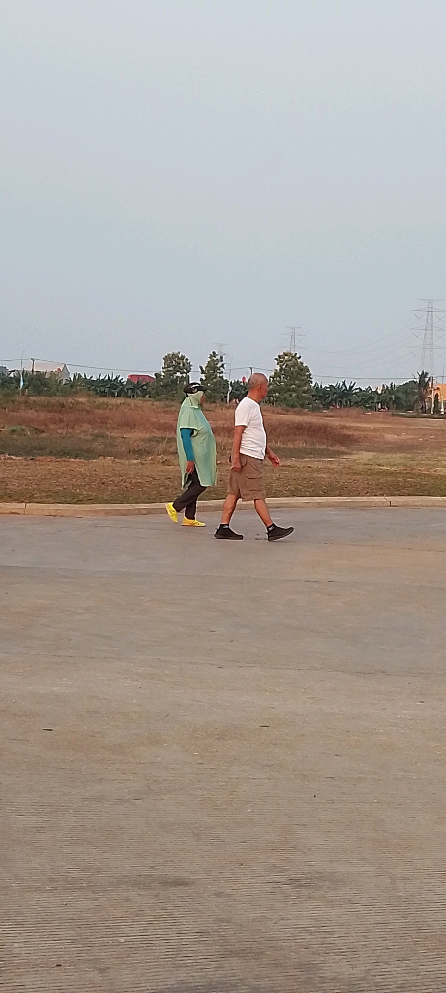 Two individuals are walking on a paved surface in Satriamekar, Indonesia. The person on the left is wearing a light green hooded garment over a blue long-sleeved shirt and black pants, with yellow shoes. This person also appears to be wearing a mask or covering over their face. The person on the right is an adult male wearing a white short-sleeved t-shirt, beige cargo shorts, and black athletic shoes. They are walking side-by-side, facing towards the right of the frame. In the background, there is a dry, grassy field with scattered trees and bushes. Further in the distance, there are power lines supported by large towers and some buildings. The sky is overcast and a pale gray. The paved surface in the foreground is a light concrete color with some faint lines.