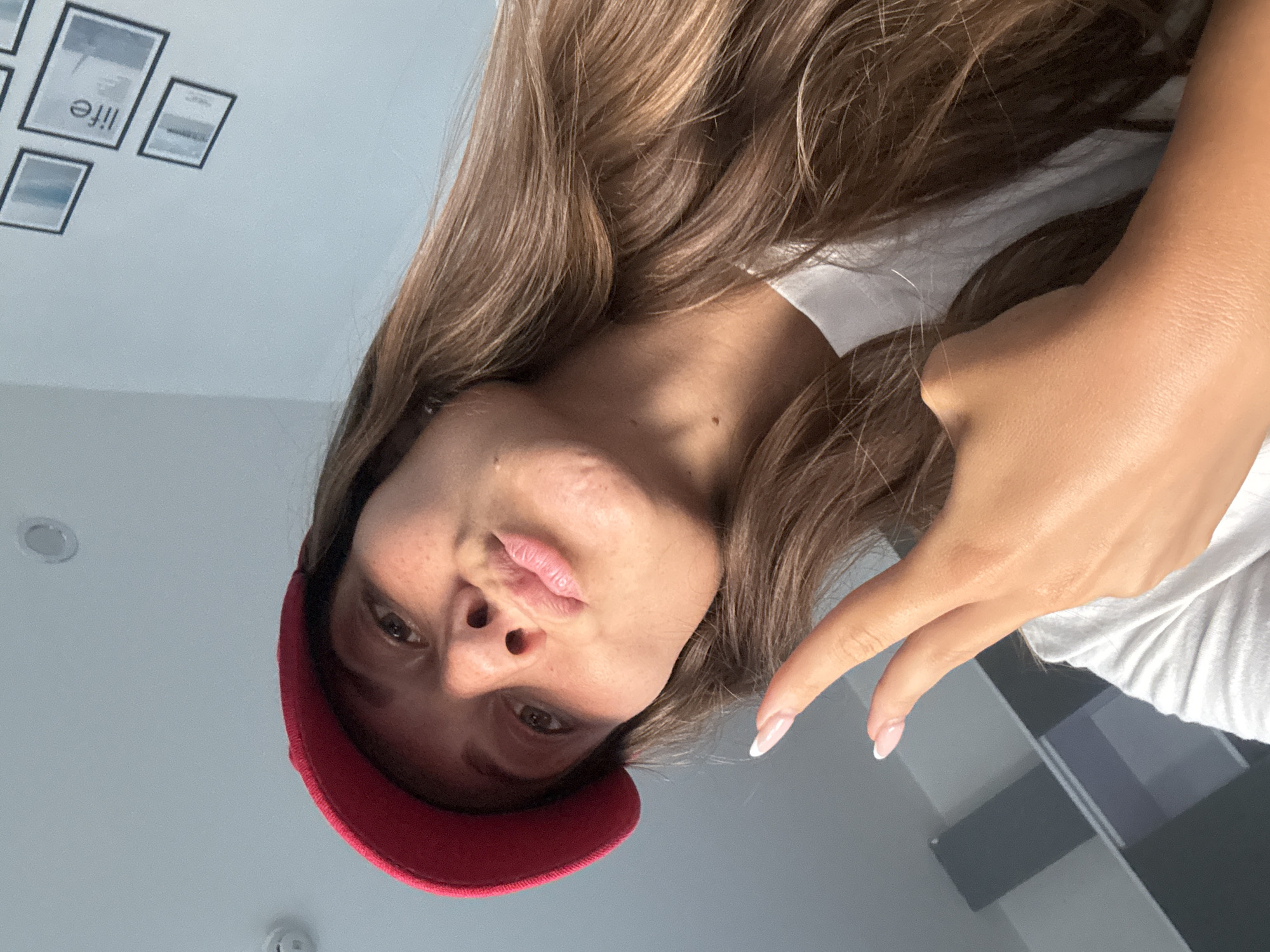 A woman with light brown hair, wearing a red baseball cap and a white t-shirt, poses for a selfie. Her face is tilted downwards towards the camera, and she is extending one hand towards the camera. The background is a white ceiling with framed pictures. The location is not discernible from this image.