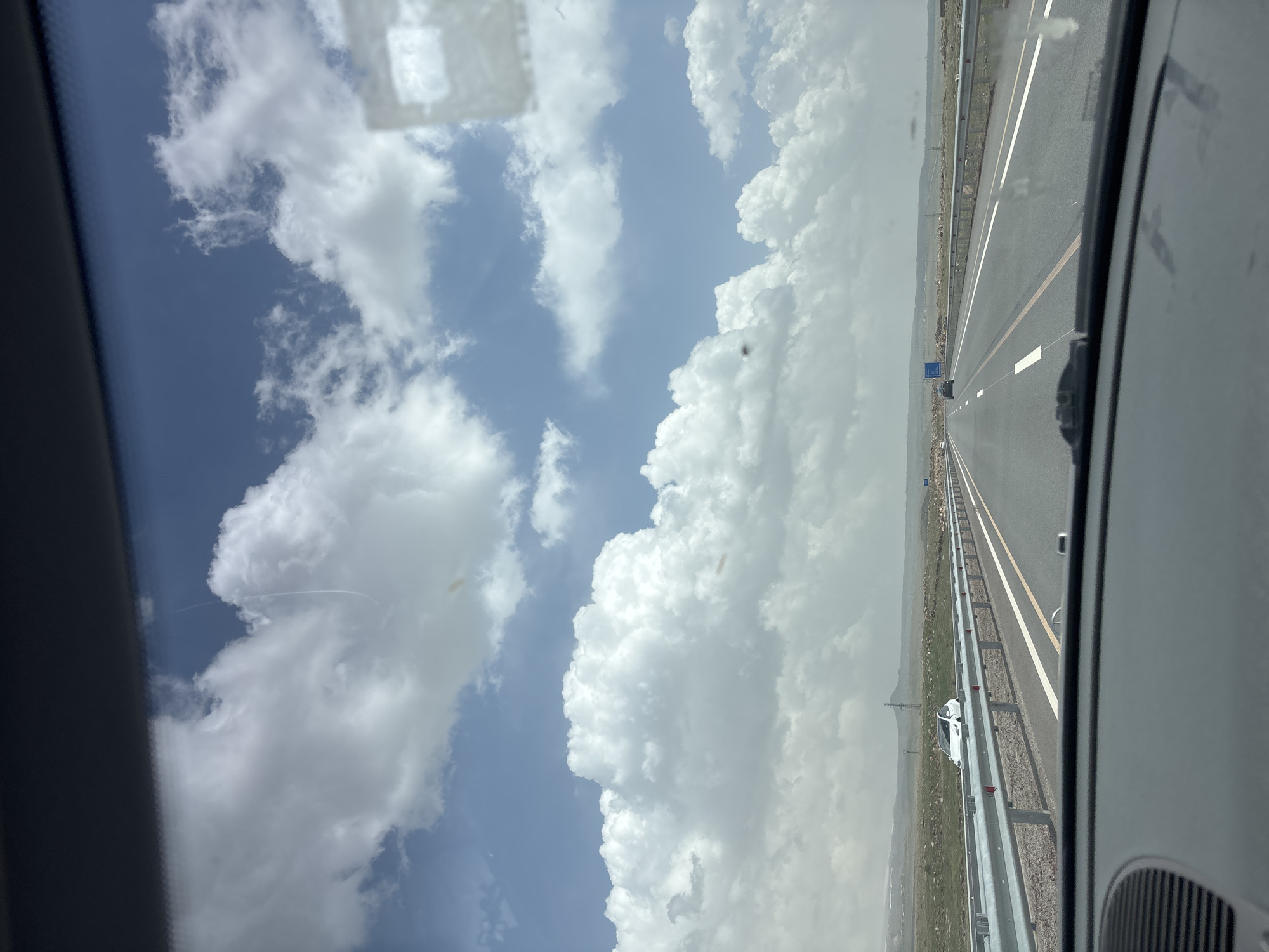 A passenger perspective of driving on a highway outside Talin, Armenia, featuring a clear blue sky with large, fluffy white clouds. The highway appears to be a multi-lane road with a yellow divider line and guardrails. A white car is visible on the side of the road. The landscape beyond the highway consists of dry, grassy terrain.