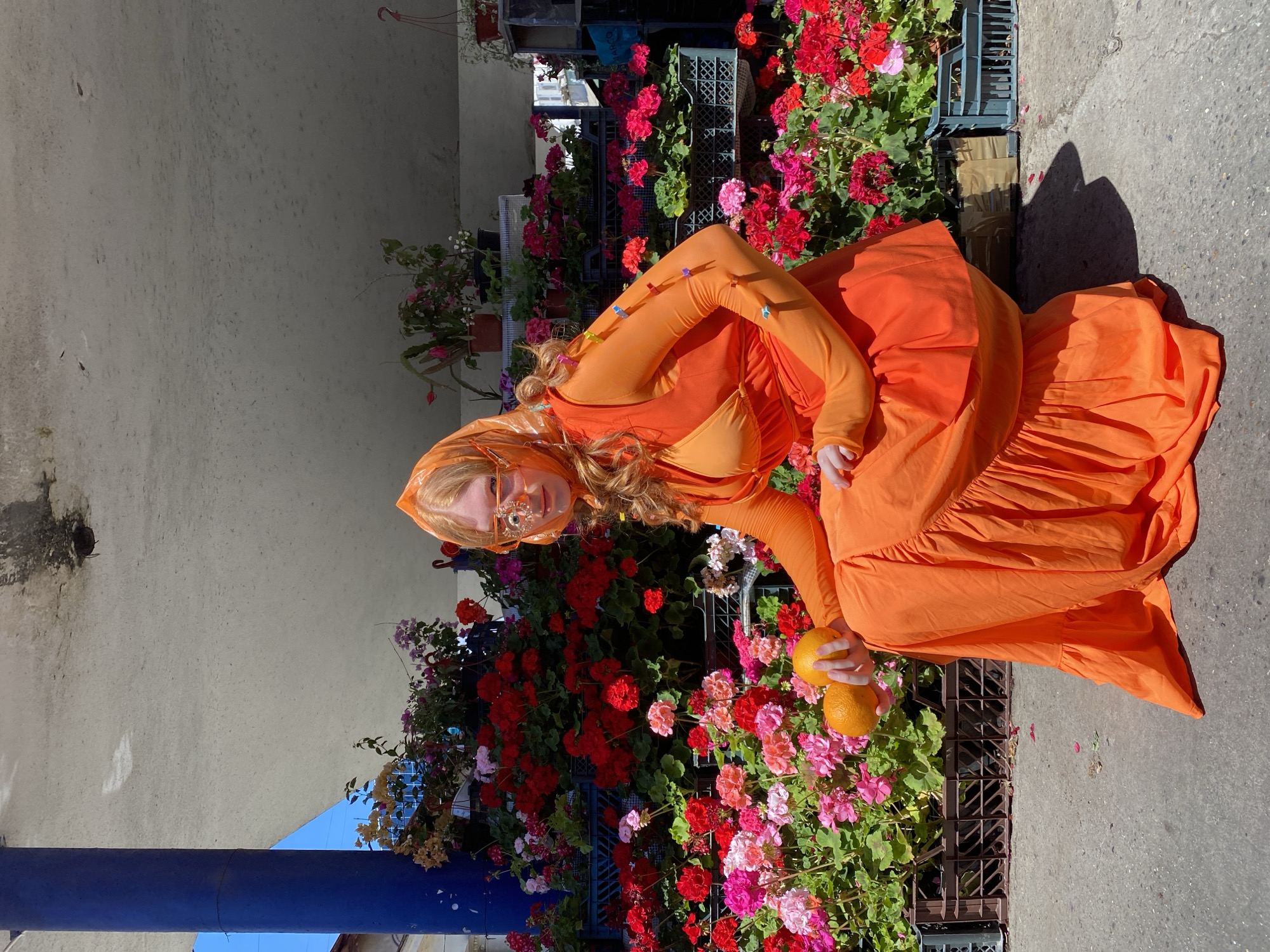 A young woman with long, reddish hair and a bright orange headscarf is crouching in front of a vibrant display of red, pink, and white geraniums. She is wearing a matching bright orange outfit, which includes a long-sleeved top and a flowing skirt. Her eyes are obscured by large, decorative eyeglasses. In her hands, she holds two oranges. The scene appears to be an outdoor market, possibly in Sochi, Russia, given the style of the stalls and the surrounding architecture. The ground is paved with concrete, and shadows suggest a sunny day. The geraniums are arranged in plastic crates, creating a colorful backdrop.