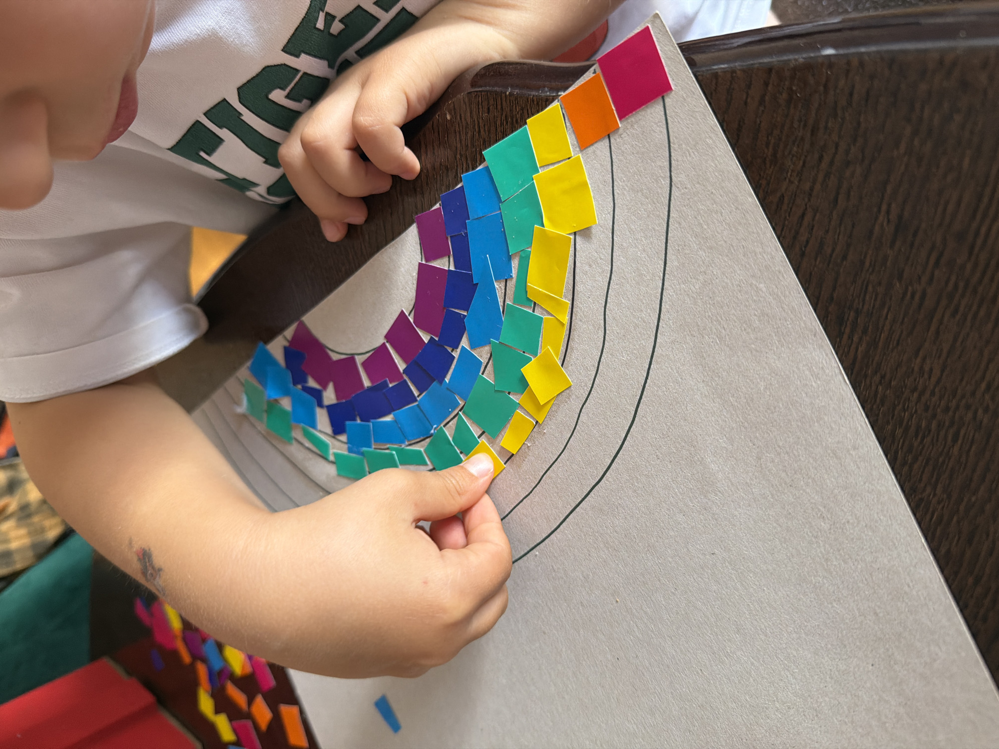 A child is making a rainbow mosaic on a gray piece of paper. The mosaic is composed of small, square pieces of paper in various colors, including pink, orange, yellow, green, blue, and purple. The child is holding a yellow piece of paper in their right hand and is placing it on the mosaic. The child is wearing a white T-shirt with green lettering. The paper is resting on a dark brown wooden surface. There is a small pile of colorful paper squares in the lower left corner of the image. The exact location is unknown.