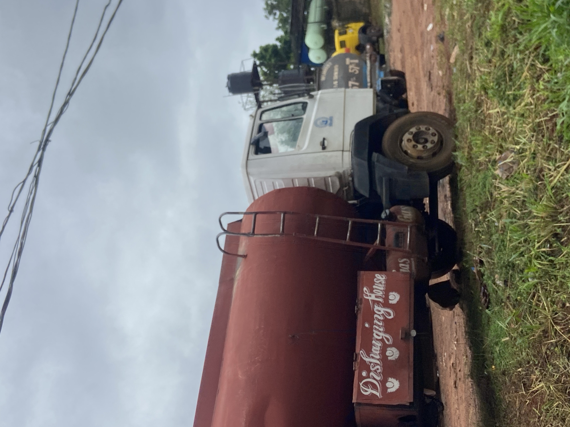 In Nkpor, Nigeria, an overcast sky looms over a scene dominated by large vehicles. The foreground is characterized by rough, reddish-brown dirt and patchy green grass. A large, red tanker truck, likely a fuel or water transport, is prominently featured on the left. It has a ladder attached to its side and writing on its lower panel that reads "Dishcharging House" and "Gas." To the right of this truck, a white truck cab is visible, with a dark-colored tanker trailer behind it, also bearing text. Further in the background, a small yellow truck is parked. The environment suggests an industrial or storage area, possibly a depot, with a somewhat unkempt appearance. Overhead, power lines crisscross the sky.