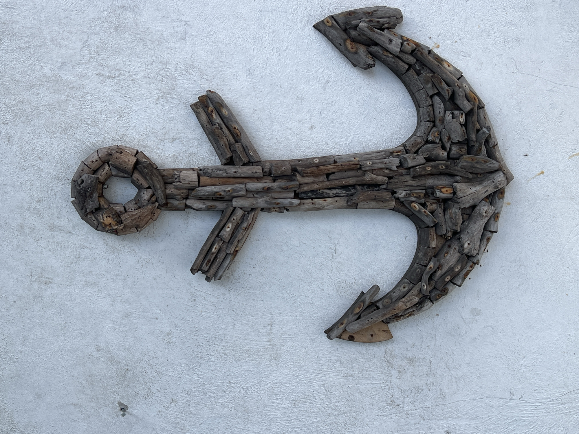 The image captures a large, decorative anchor art piece meticulously crafted from numerous small, weathered pieces of wood, prominently displayed against a textured white wall. The anchor’s traditional form, including its stock, shank, and flukes, is constructed entirely from these irregularly shaped wooden segments, giving it a rustic, driftwood-like appearance. Small nails or pins are visible throughout the piece, fastening the individual wooden elements together. The wood displays a natural variation in tone, ranging from light greys to deeper browns, indicative of its aged and possibly repurposed nature.
The background is a roughly textured, stucco-like wall, painted in a bright off-white or light grey color. This neutral backdrop allows the intricate details and varied textures of the wooden anchor to stand out clearly. The lighting is soft and even, suggesting either an overcast day or diffused indoor illumination, which minimizes harsh shadows and enhances the tactile quality of both the wood and the wall. No human activity is depicted; the anchor is a stationary decorative object. There is no visible text in the image. This scene, set in the Ubud District, Indonesia, evokes a calm, handcrafted, and subtly nautical aesthetic.
