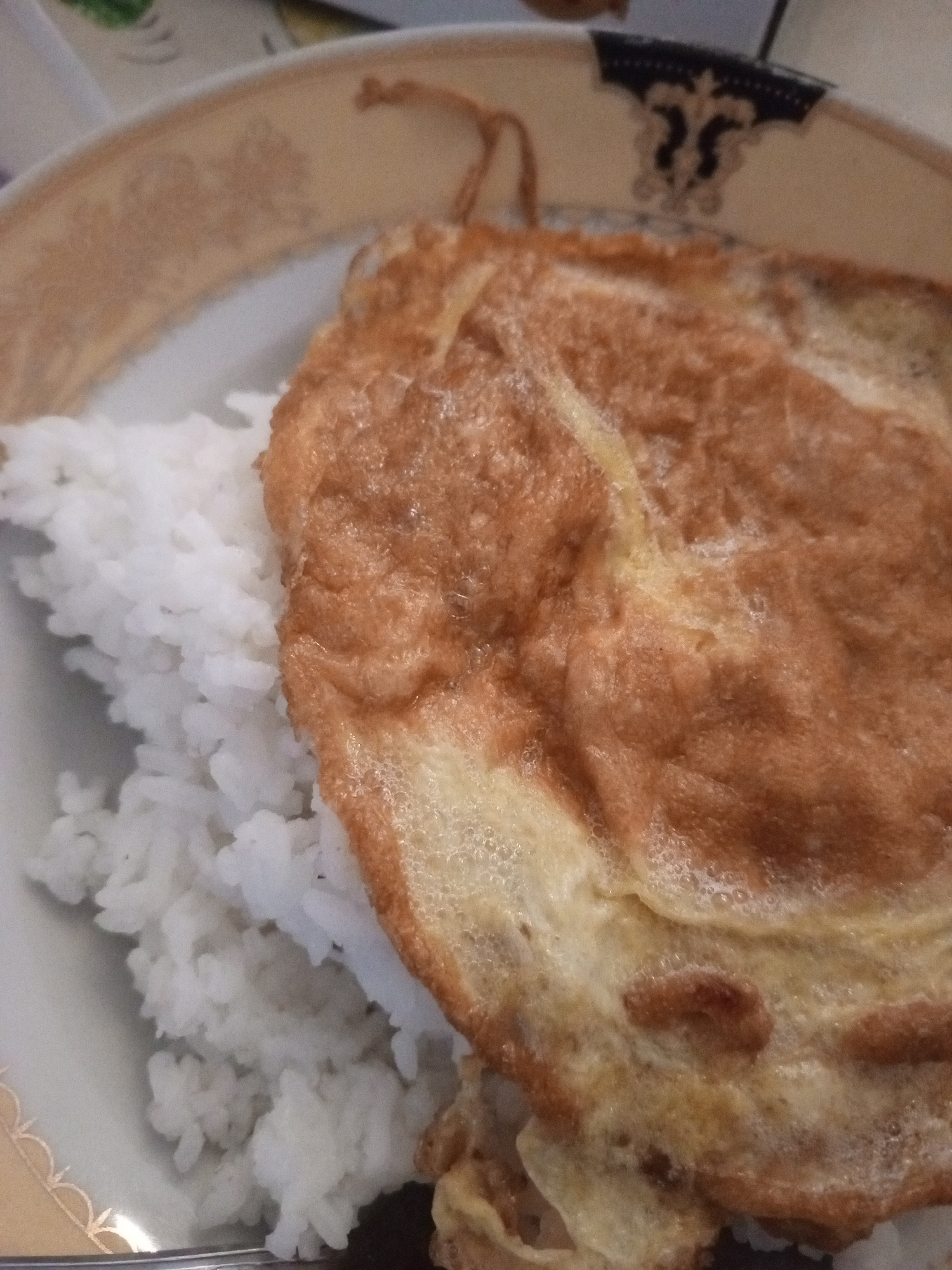 A close-up, slightly overhead shot shows a plate of white rice topped with a golden-brown omelet. The rice is fluffy and piled high on the left side of the plate, while the omelet, which appears to be cooked evenly with some slight browning and a bubbly texture, covers most of the right side. The plate itself is white with a decorative gold trim and a darker blue and black pattern around the rim. The image is taken in Mvera Mission, Malawi, where this simple yet appetizing meal is being served.