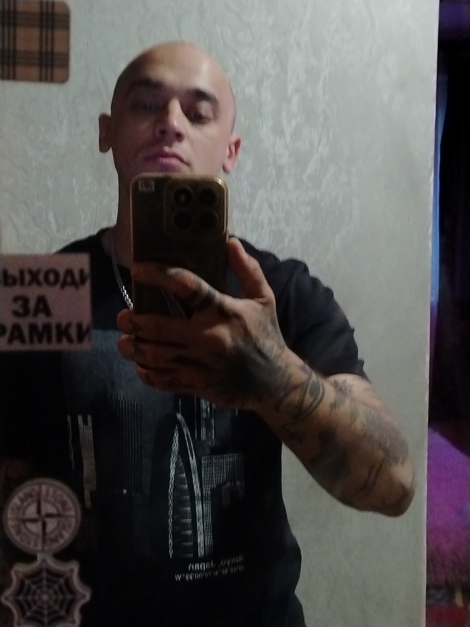 In an indoor setting, a bald man is taking a mirror selfie. He is holding a smartphone in his right hand, angled to capture his reflection. He is wearing a dark t-shirt with a graphic design of buildings. On the left side of his chest, there are two circular emblems attached to his shirt; one resembles a compass rose, and the other is a spiderweb design. His left arm is covered in tattoos. In the reflection of the mirror behind him, on the left side, a sign with Russian text is visible, reading "ВЫХОДЫ ЗА РАМКИ" which translates to "EXIT OUTSIDE THE FRAMES." The background appears to be a textured wall.