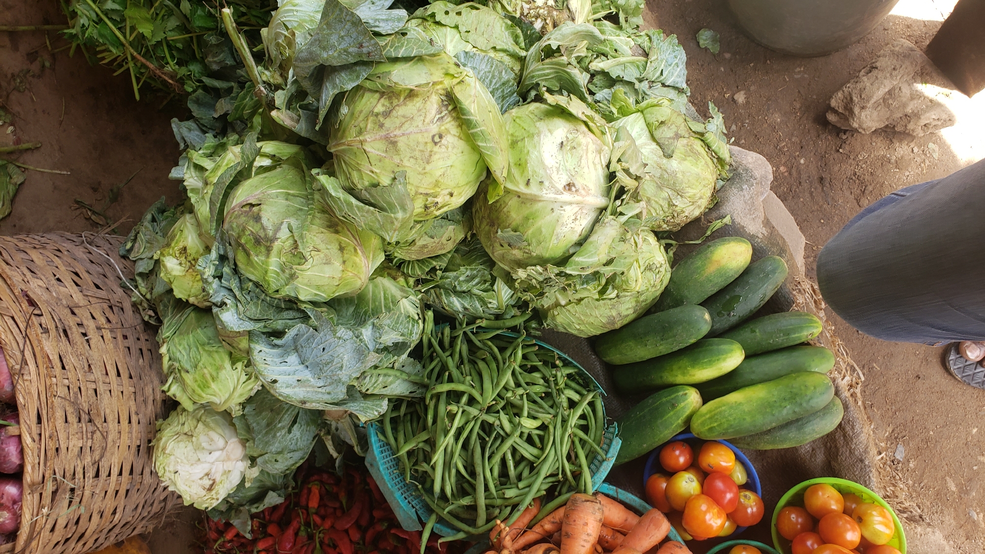The image depicts a vibrant produce display in Sabon Tasha, Nigeria. A large woven basket containing red onions sits alongside a pile of green cabbages. In front of the cabbages, a blue basket overflows with green beans. Small plastic bowls hold orange carrots and a mix of red and orange tomatoes. A row of dark green cucumbers lies to the right of the vegetables. A portion of a person's leg, clad in dark blue denim, is visible in the upper right corner of the image. The scene is set on a patch of reddish-brown soil. The lighting suggests an outdoor, daylight setting.