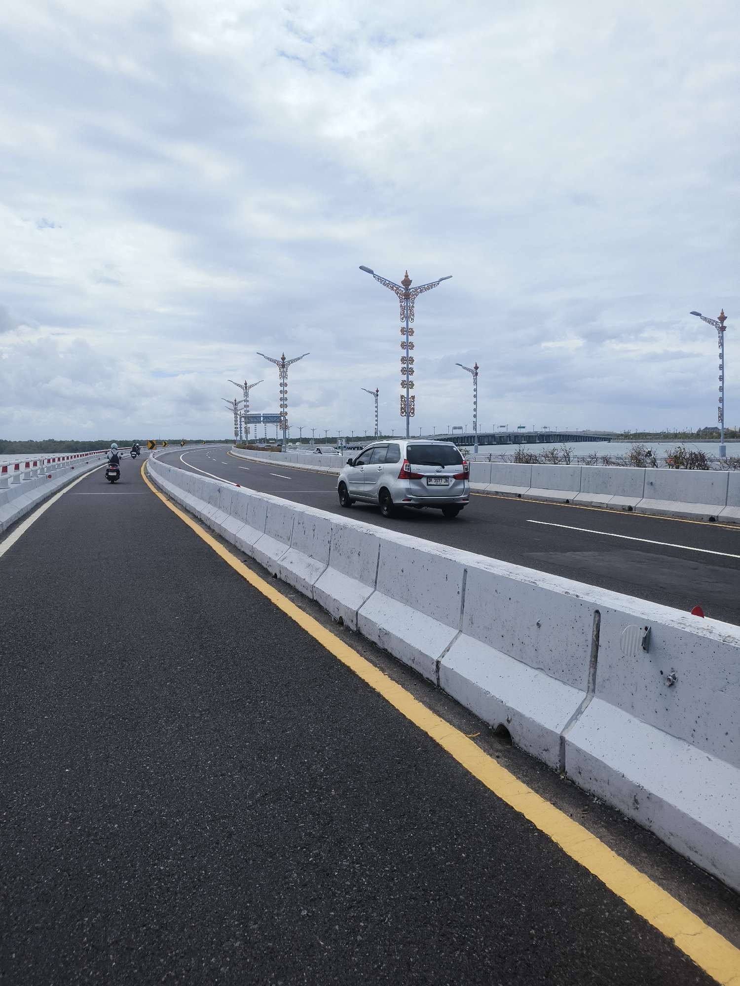 An outdoor, eye-level, wide shot captures a view of the Bali Mandara Toll Road in South Denpasar, Indonesia, under an overcast sky. The dark asphalt road dominates the foreground, with a bright yellow line marking the lane. Concrete barrier walls line the right side of the road, and on the left, white and red barriers indicate the edge of the road. Several motorcycles are traveling in the distance on the left lane, and a silver minivan is moving forward on the right lane. Tall, ornate streetlights with decorative elements and lights stand along the right side of the toll road, interspersed with modern signposts. The background shows a body of water and a distant bridge. The overall atmosphere is one of an active, modern infrastructure project.