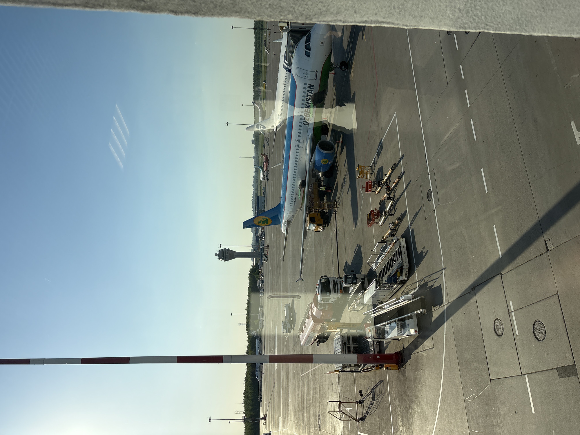 The image shows an airport apron, viewed from a window. Two Uzbekistan Airways planes are visible, along with ground service equipment. In the background is an air traffic control tower. The view is presumably from Pulkovo Airport in St. Petersburg, Russia. A red and white striped boom barrier is visible in the foreground.