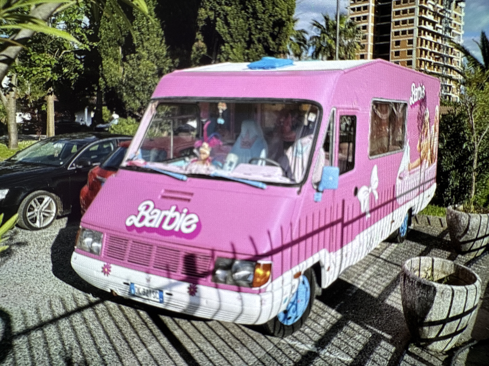 A pink Barbie campervan is parked in an outdoor area in the city of Budva, Montenegro, as identifiable from the vehicle registration plate. The campervan has a white lower body and light blue wheels. The Barbie logo is displayed on the front of the vehicle. Through the front windshield, one can see dolls and other objects. The vehicle is parked on pavement with shadows cast from nearby foliage. A black car is parked to the left, and a red car is partially visible behind the campervan. In the background, there is a partially constructed high-rise building and a variety of trees and shrubs. Two decorative planters resembling barrels are visible on the right side of the image.
