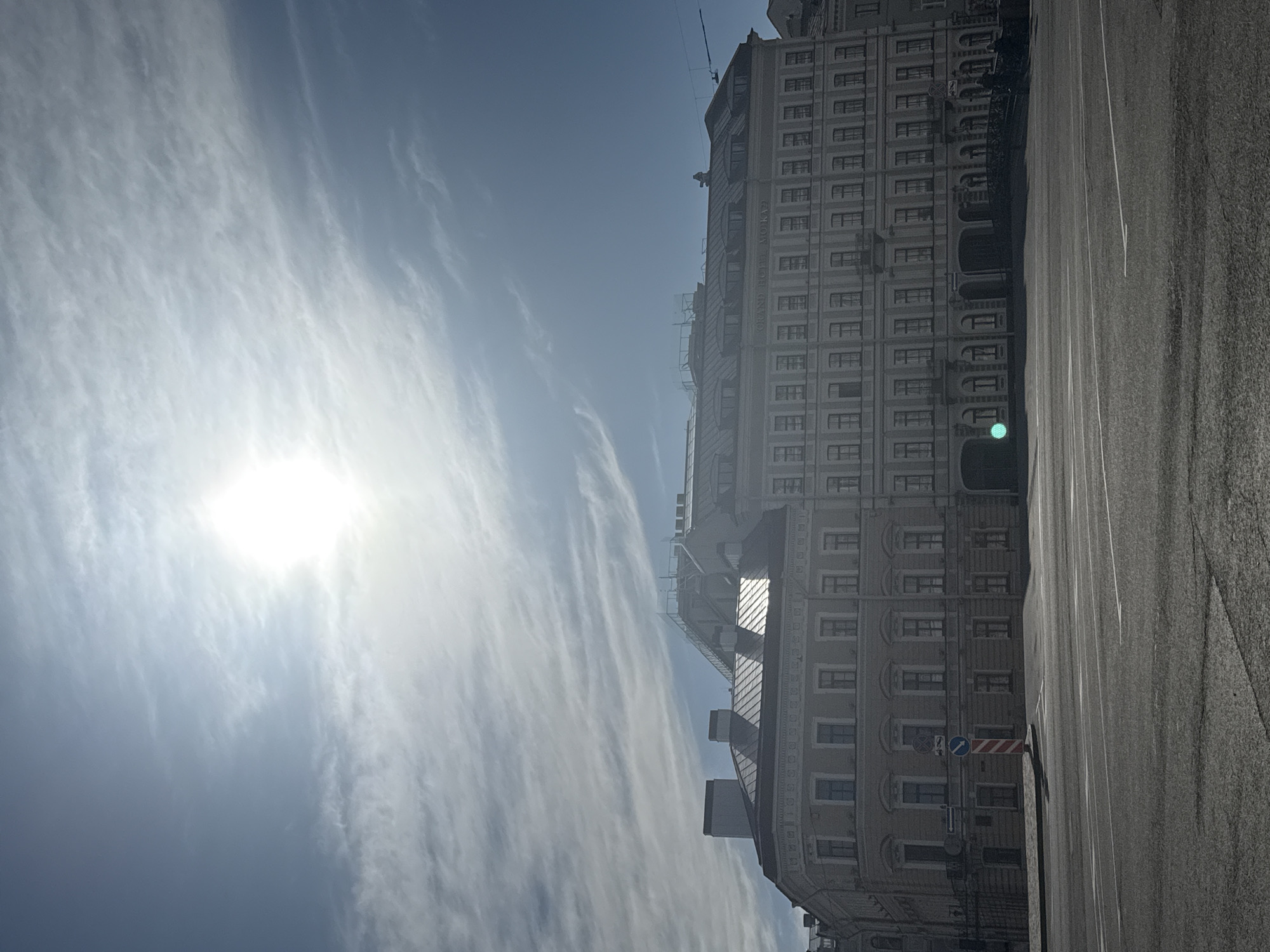 The image shows a view in Saint Petersburg, Russia, featuring the Grand Hotel Europe. The building, a multi-story structure with numerous windows and architectural details, occupies a significant portion of the right side of the frame. A paved road stretches alongside the building. The sky, a bright blue with wispy, cirrus-like clouds, fills the majority of the left side, with the sun a hazy point of light.