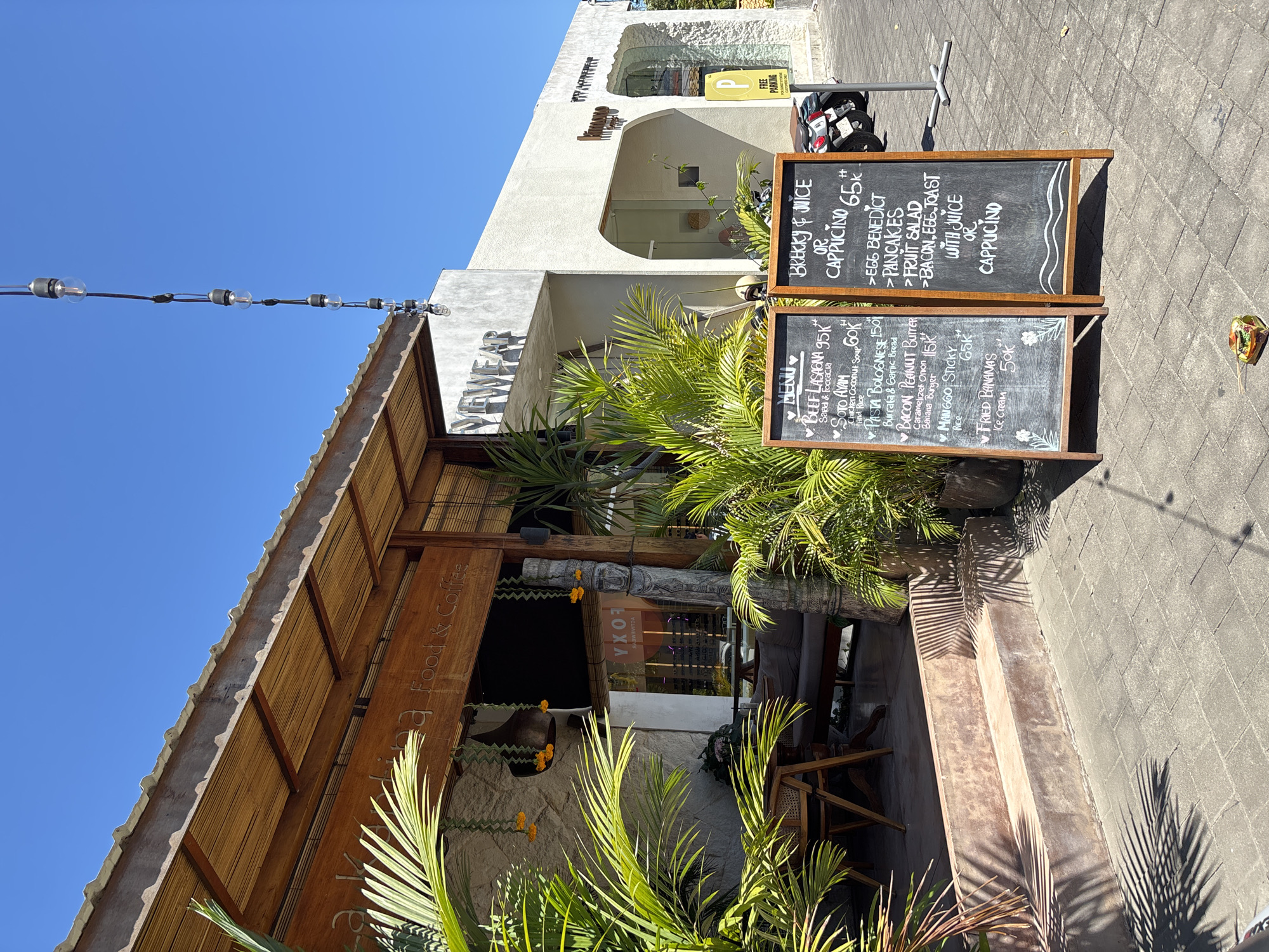 The image shows the exterior of "Dewear," a food and coffee establishment in Pecatu, Indonesia. The building has a bamboo-covered roof and a stone facade. A vertical sign displays "a Food & Coffee" next to the restaurant's name. Palm plants surround the entrance. Two blackboards display menus. One blackboard advertises "Brecky & Juice or Cappuccino 65k++" followed by a list of breakfast items. The second displays menu items such as beef lasagna (95k++), soto ayam (60k++), pasta bolognese (150k++), and other dishes with prices. A white building is to the right, displaying the sign for the parking lot. The sky is blue. The foreground shows part of a paved sidewalk.