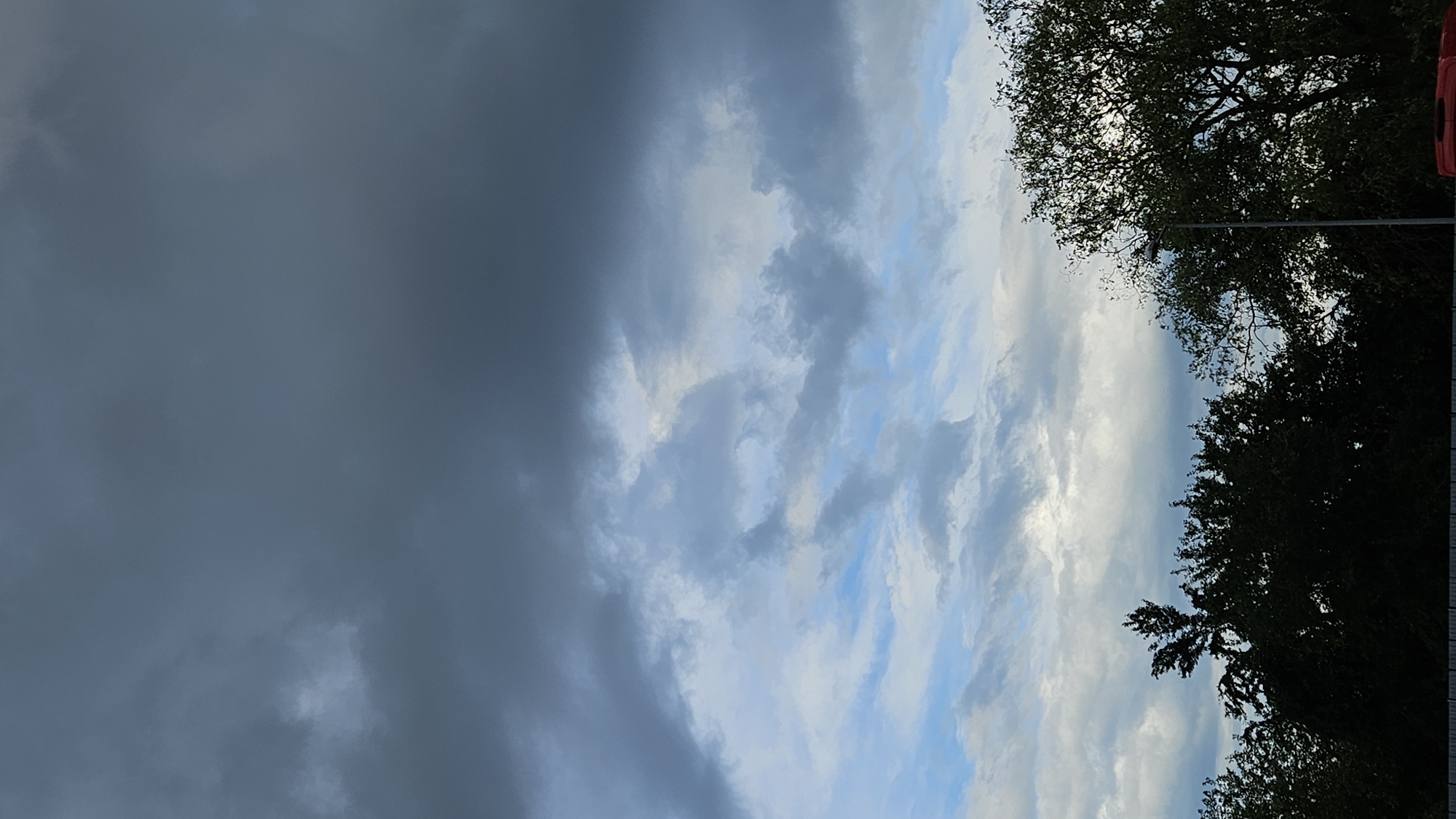 The image depicts a cloudy sky over a line of trees. The sky is a mix of dark gray storm clouds in the upper portion, transitioning to lighter gray and patches of blue sky with lighter, scattered clouds below. The trees are silhouetted against the sky, appearing dark green and black, with some branches visible. One street light can be seen amongst the trees on the right side of the frame. The bottom edge of the image shows a darker, horizontal element, possibly a fence or the roof of a vehicle, with a hint of red visible at the very bottom right, suggesting a vehicle. There are no people visible in the image. The scene is likely an outdoor environment, possibly a park or the edge of a residential area, given the presence of trees and a street light. The specific location in Prešov, Slovakia is not discernible from the image itself, as there are no identifying landmarks or signs.