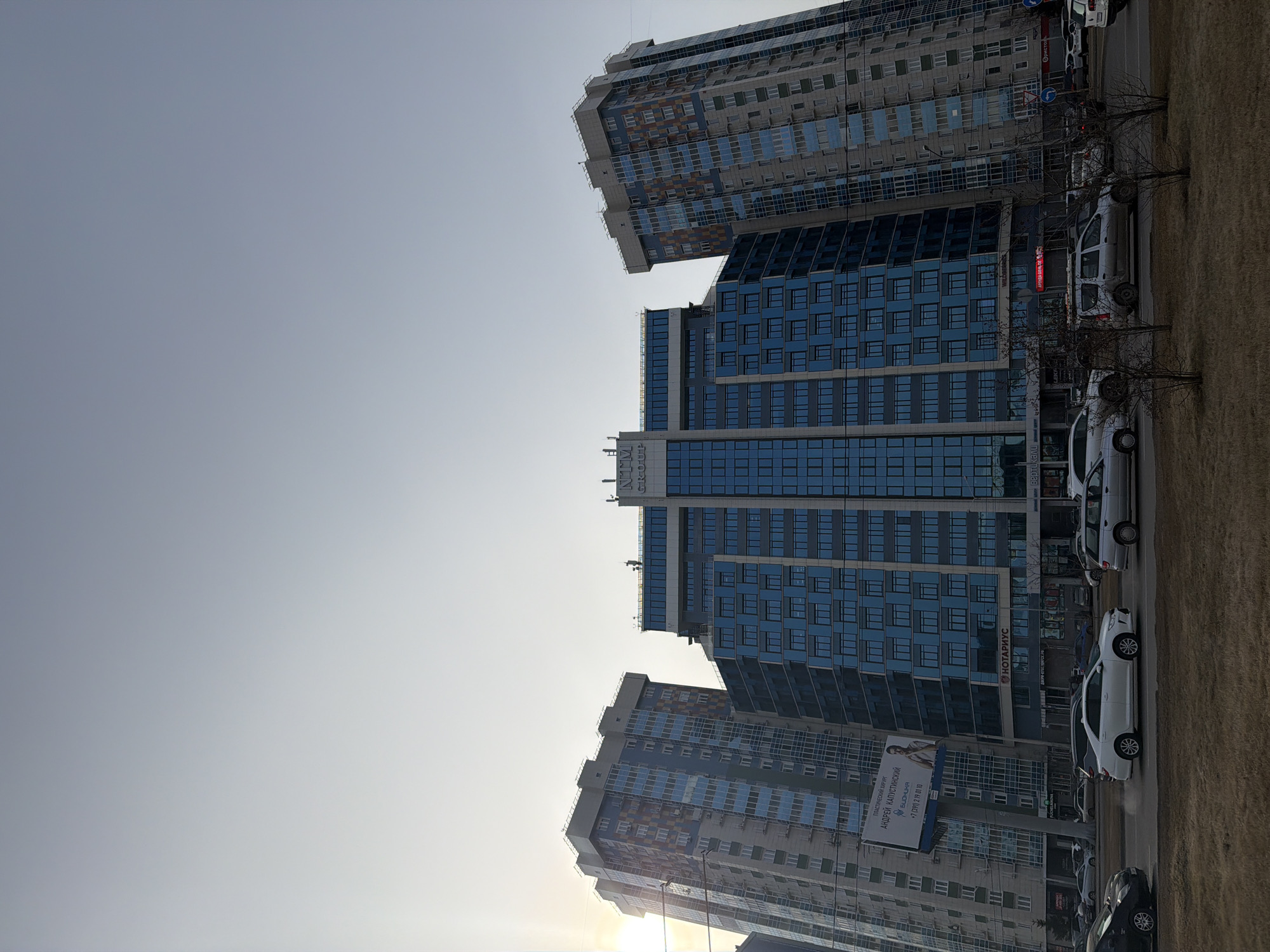 The image depicts a street scene in Krasnoyarsk, Russia. Three modern high-rise buildings dominate the view. The central building features a sign with "NTM GROUP" written on it. A row of parked cars lines the street. A billboard advertising a plastic surgeon named Andrey Kapustinsky is visible. Leafless trees stand between the sidewalk and the road. The sky is pale and clear.