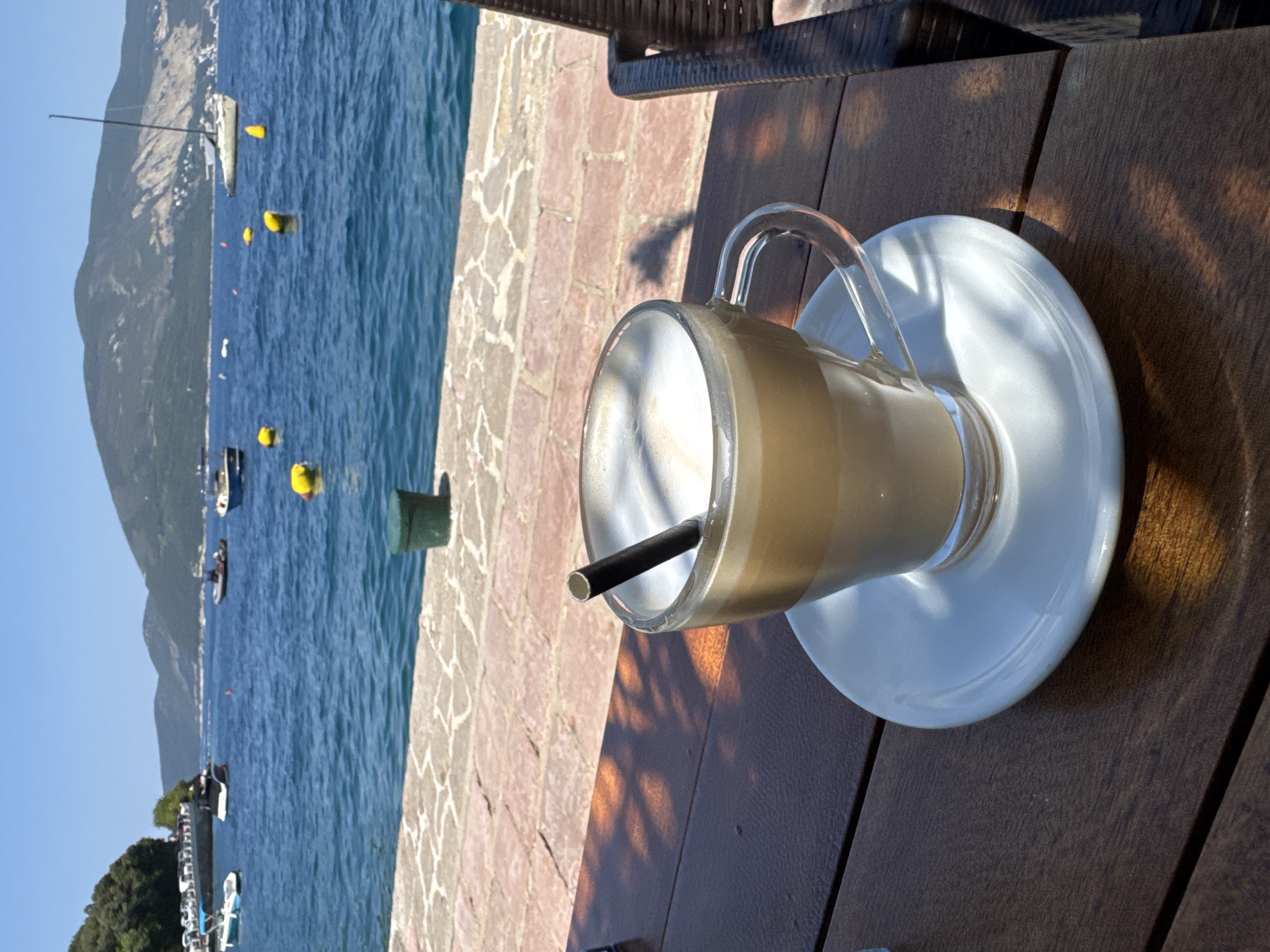 A tall glass mug filled with a frothy, light brown beverage, likely a coffee drink, sits on a white saucer atop a wooden table. A black straw extends from the mug. The mug is made of clear glass with a handle. Sunlight casts dappled shadows on the table and the saucer.
The setting appears to be outdoors, with a body of water in the background, suggesting a seaside or lakeside location. The water is a vibrant blue, and several yellow buoys are visible. In the distance, a sailboat with a tall mast is moored. A mountain range with sparse vegetation lines the far side of the water. The shore is paved with stones in a mosaic pattern. The lighting indicates it is daytime, likely morning or afternoon, with clear weather. The scene evokes a sense of relaxation and enjoyment. There is no visible text in the image.